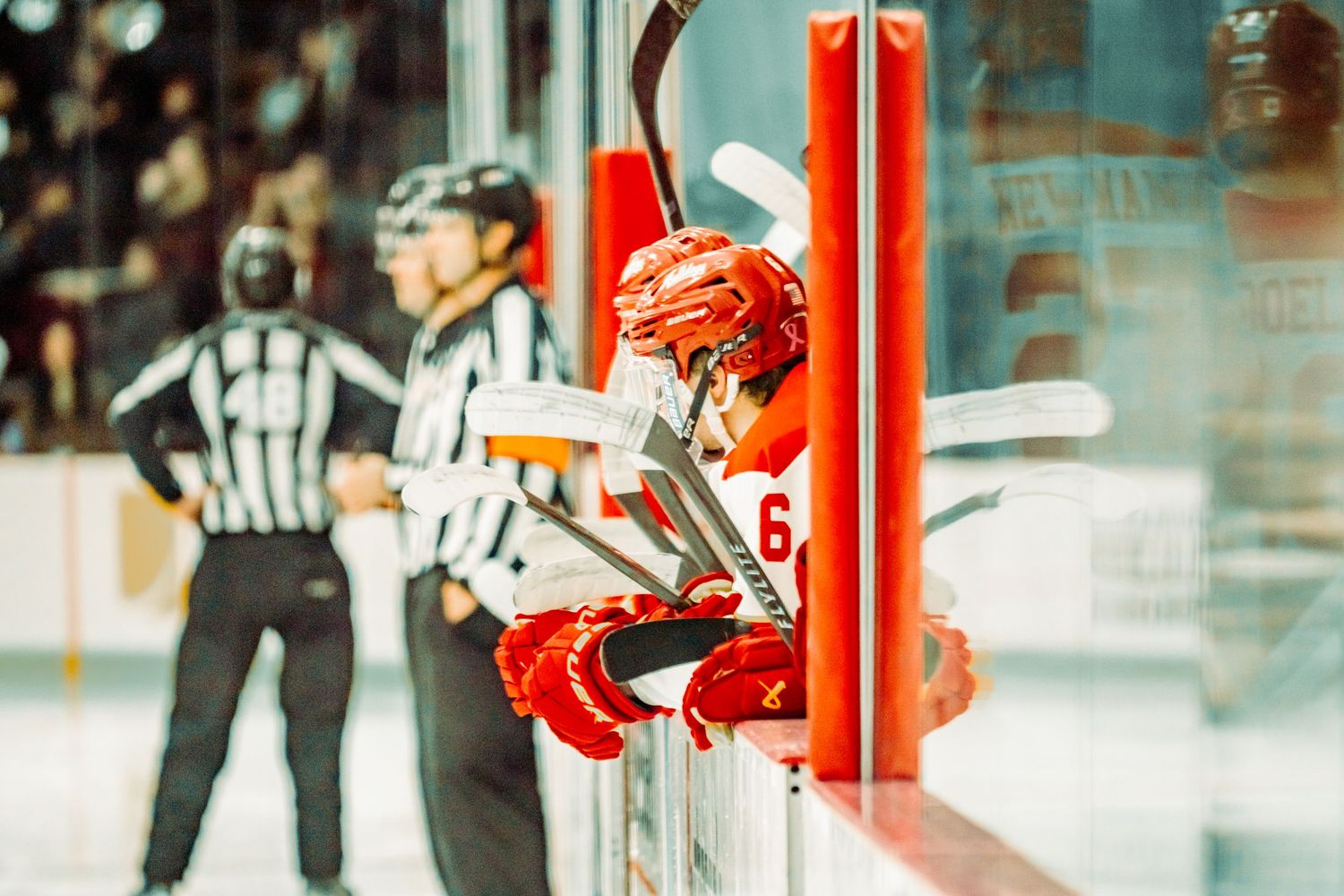 Red-helmeted hockey player in a red jersey leans on the boards while two referees in striped shirts stand on the ice behind him.