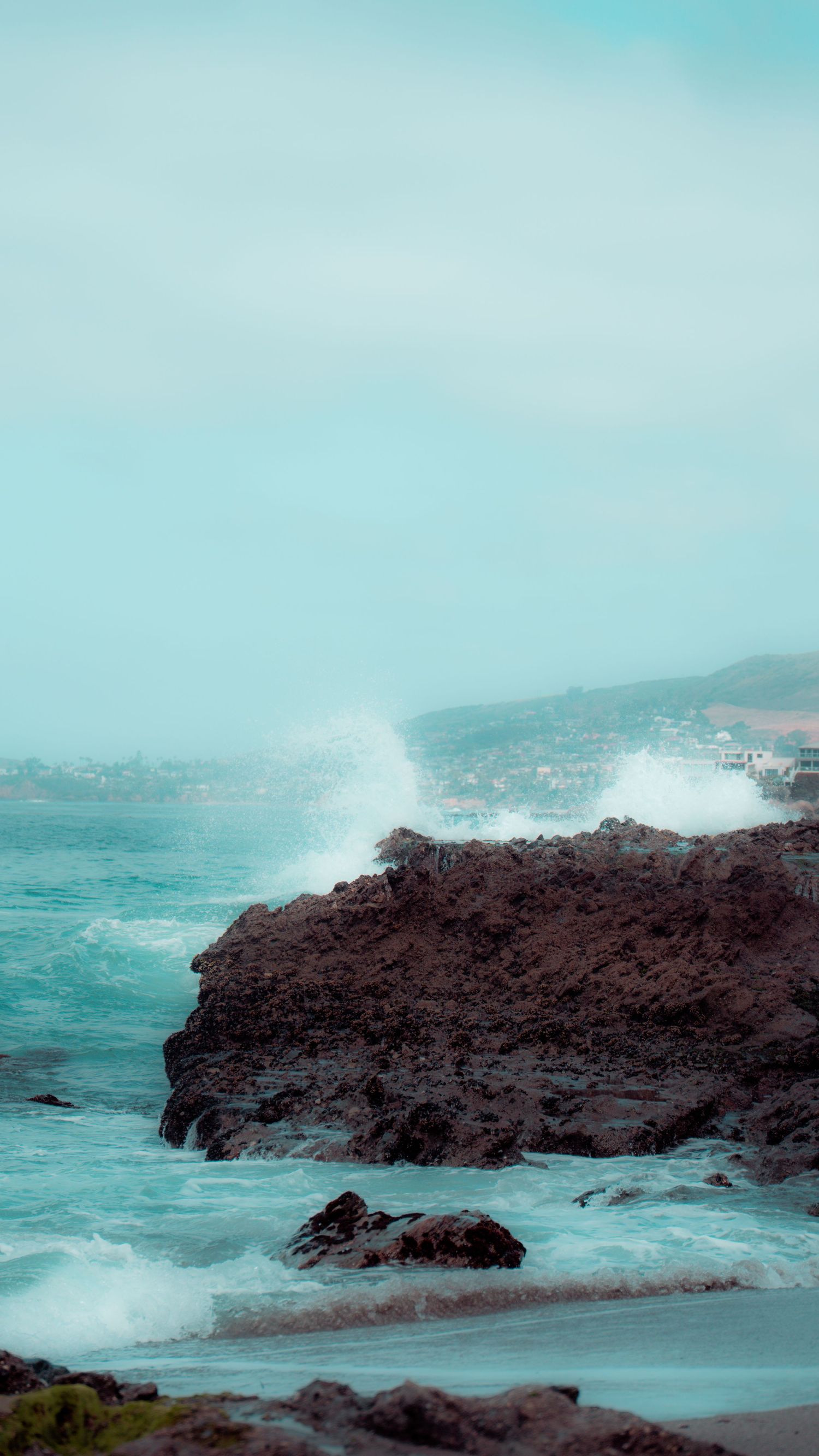 Dramatic ocean waves crash against a rugged coastal cliff as white spray erupts into the turquoise sky.