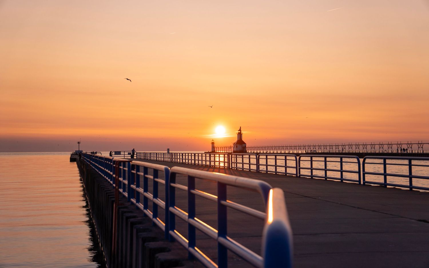 Long pier extends into Lake Michigan with lighthouse silhouette against vibrant orange sunset sky.