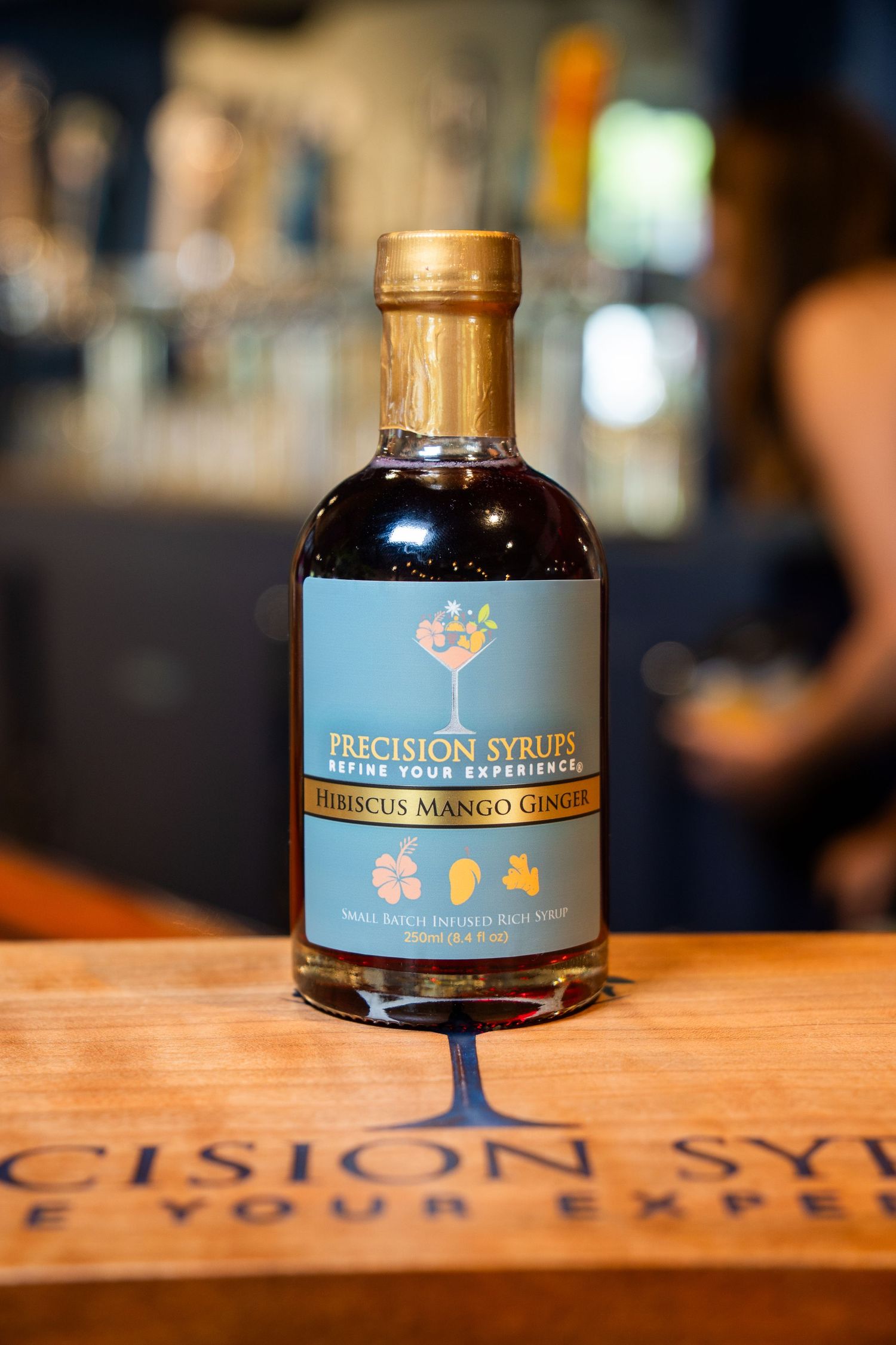 Precision Syrup's slumbering mermaid bottle displayed on branded wooden surface in bar setting.