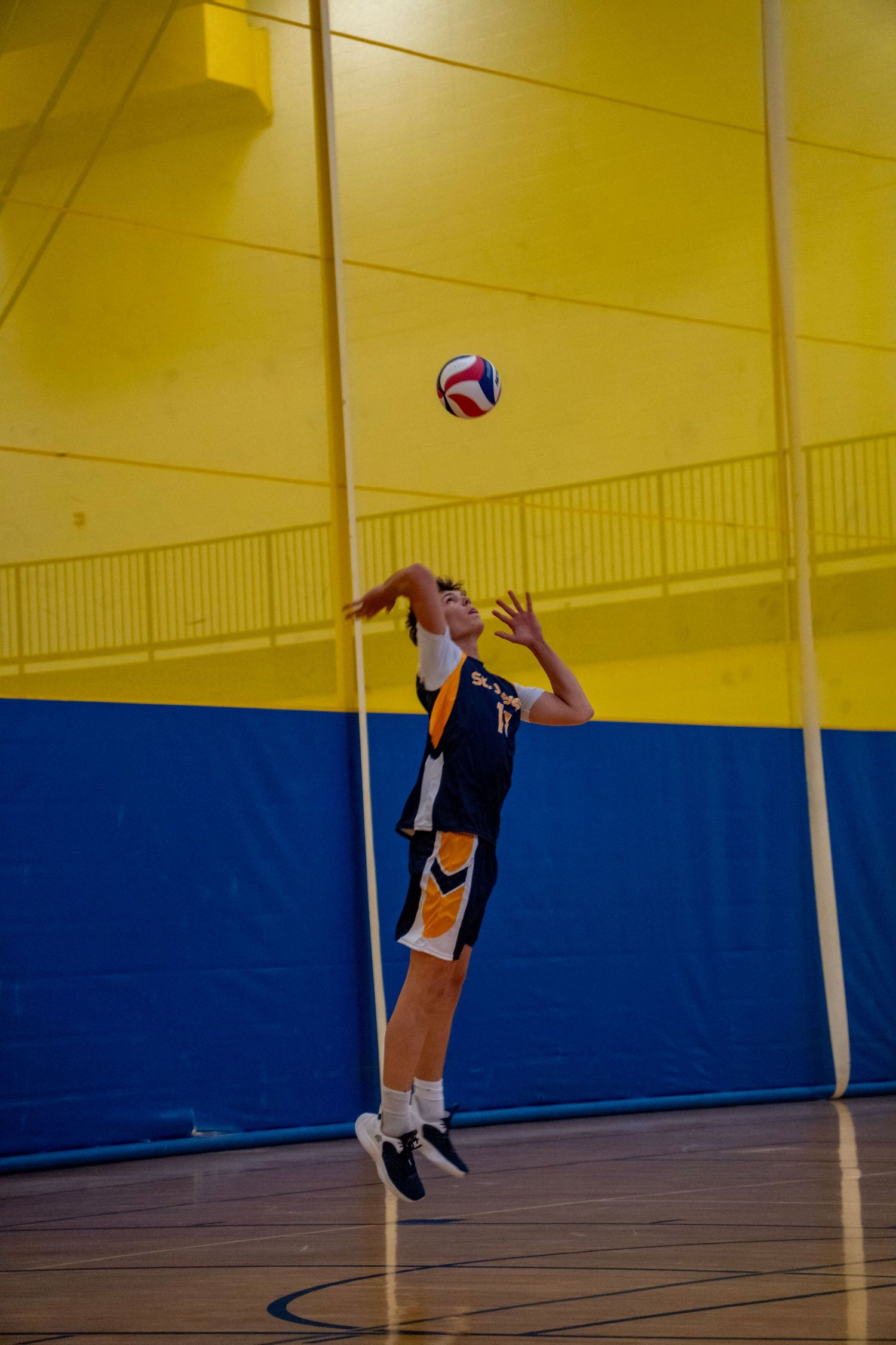 Player in navy and yellow uniform jumping to serve the ball against yellow wall background.