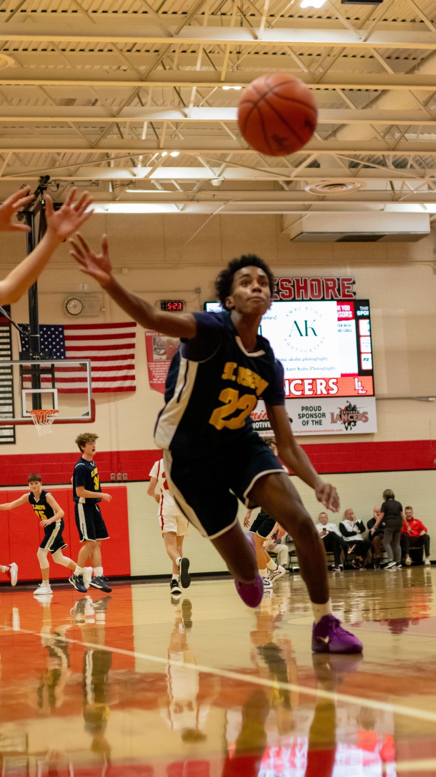 Basketball player in black jersey leaps high for layup with American flag visible in gymnasium.