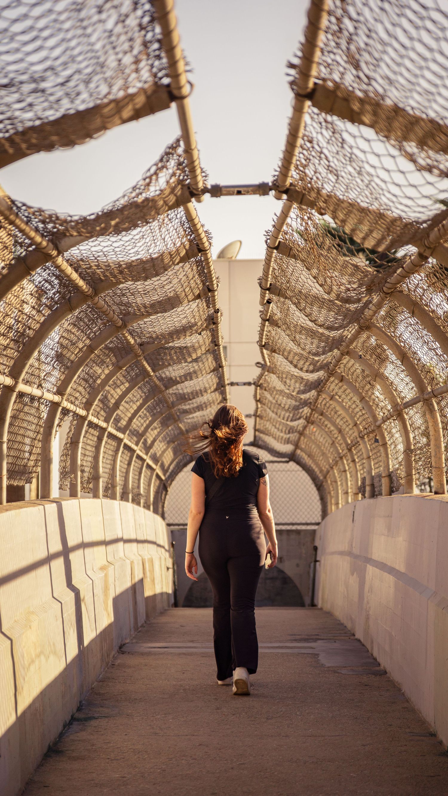 Figure walking through architectural tunnel with mesh overhead and geometric shadows.
