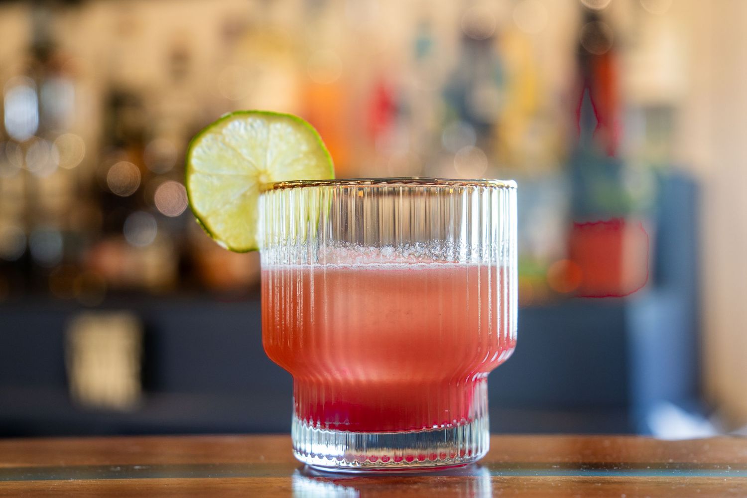 Pink craft cocktail served in ribbed glass with lime wheel garnish on wooden surface.