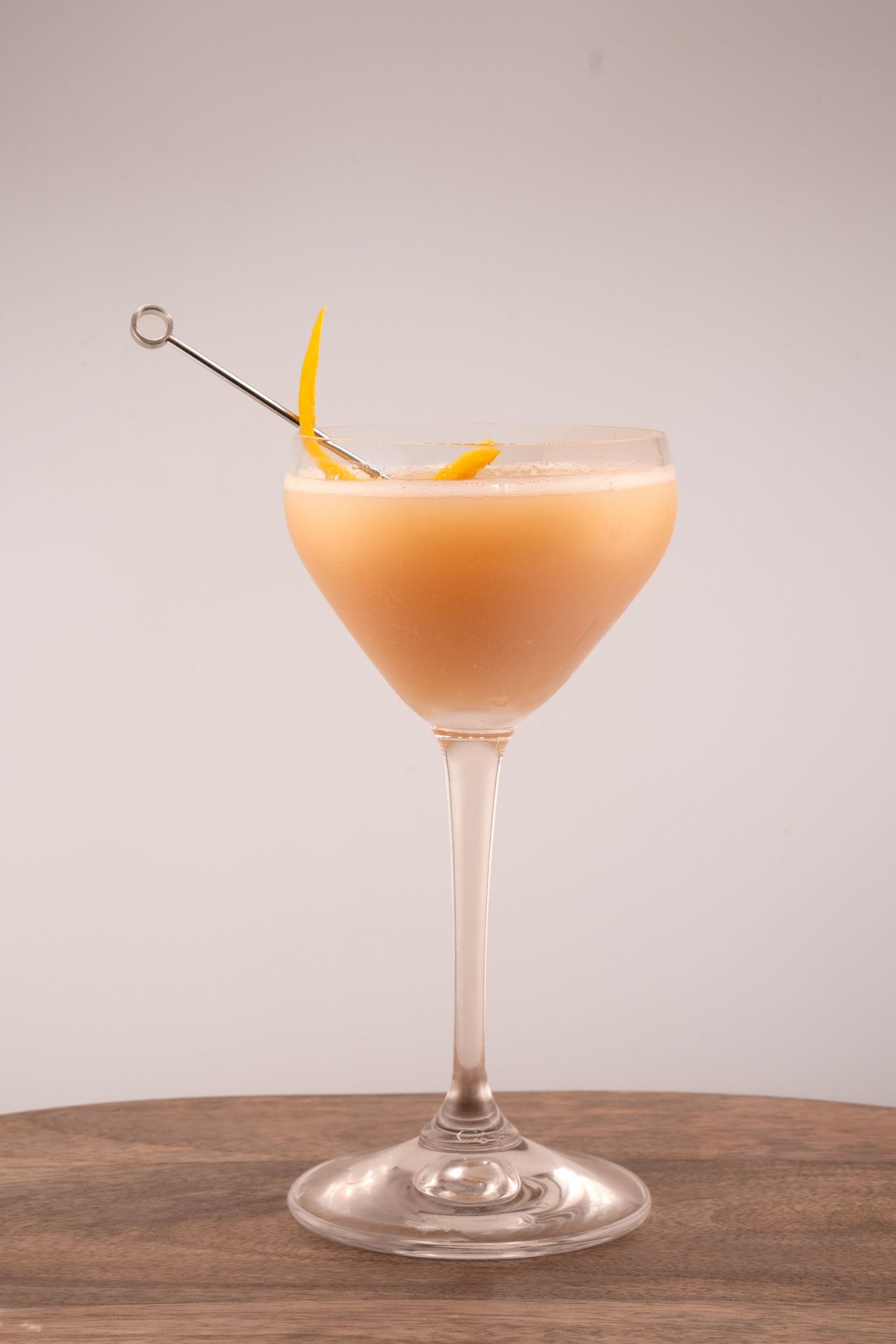 Peachy-pink cocktail in classic coupe glass garnished with orange peel and cocktail pick.