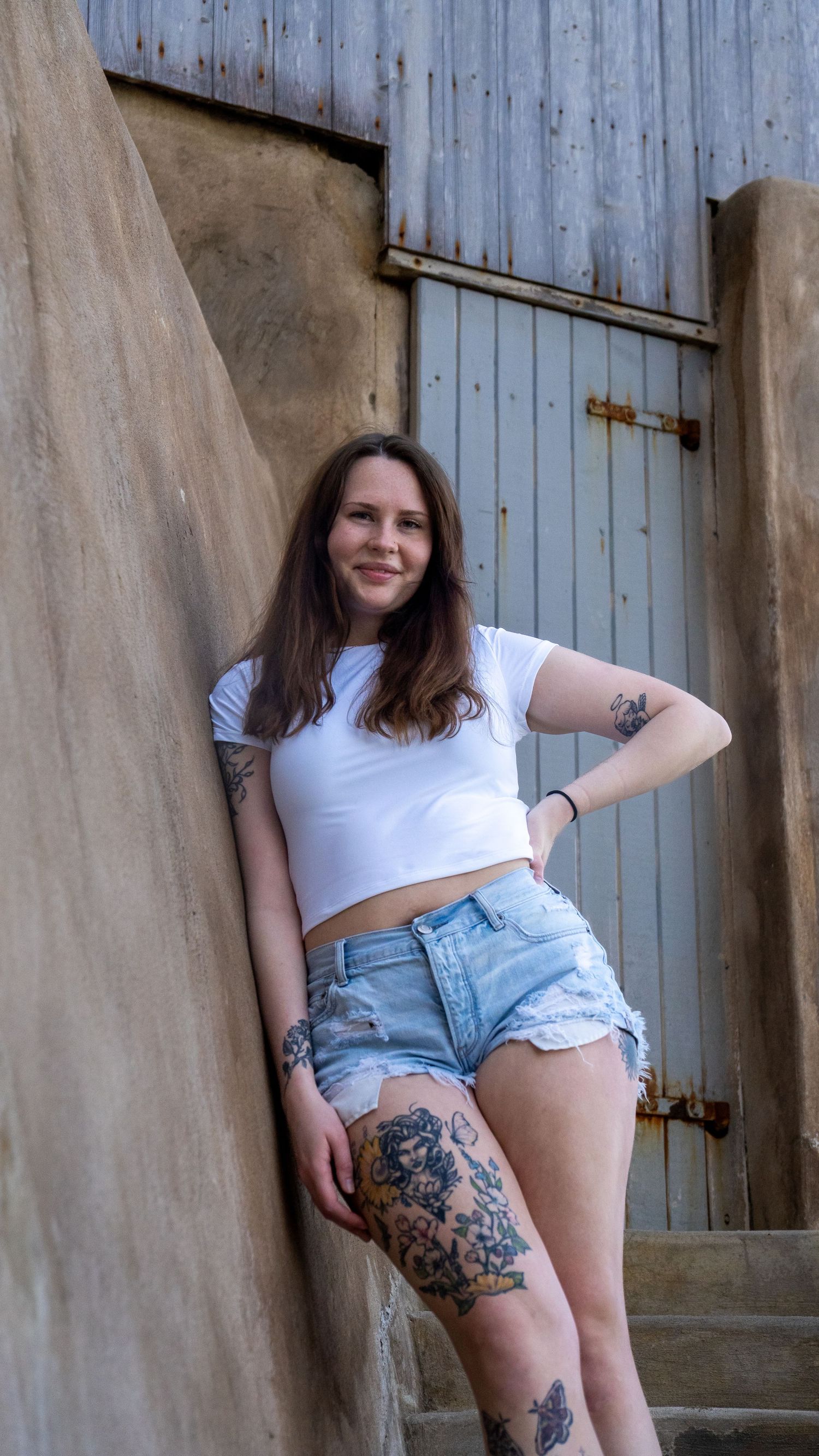 Casual summer portrait against textured wall featuring artistic tattoos and denim shorts.