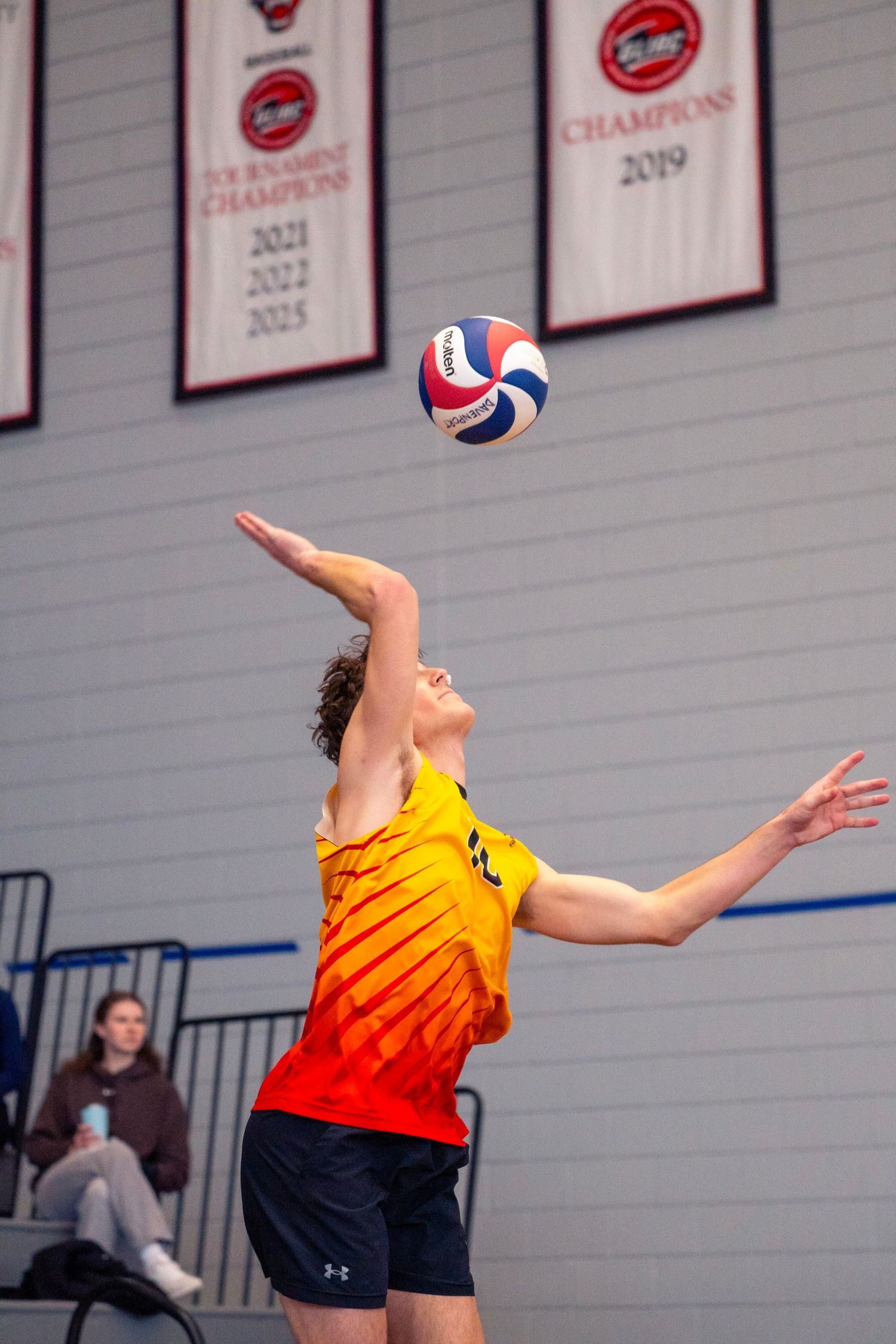 Volleyball player in orange gradient jersey jumps to serve ball during indoor match with championship banners visible.