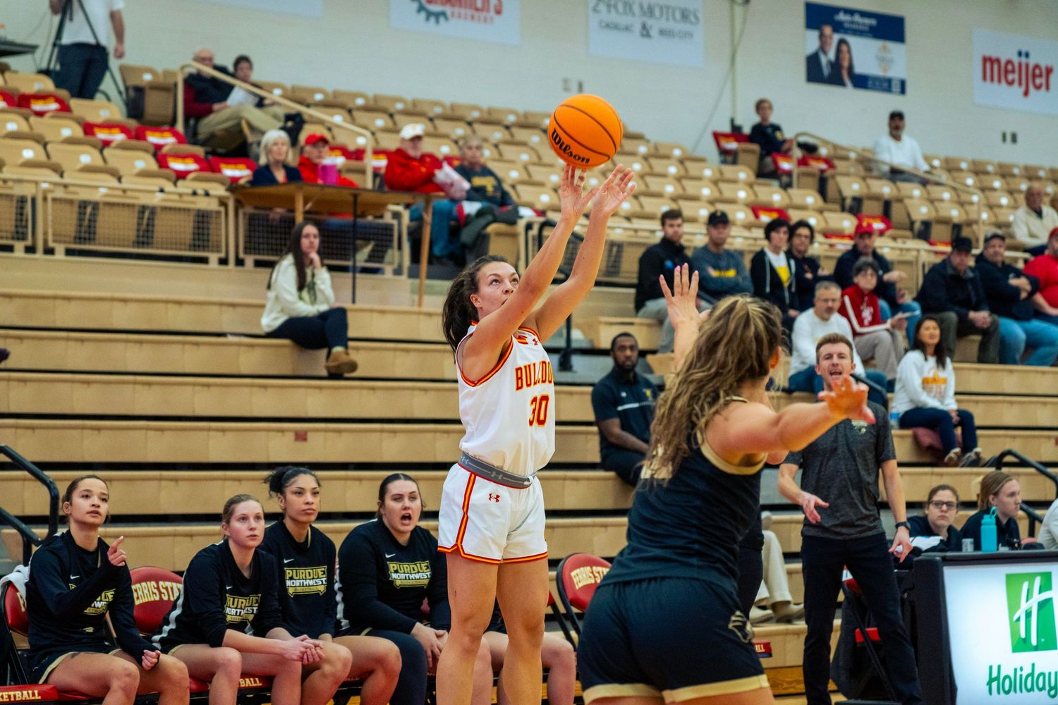 Basketball players compete for a rebound during an intense game in a gymnasium with spectators watching from bleachers.