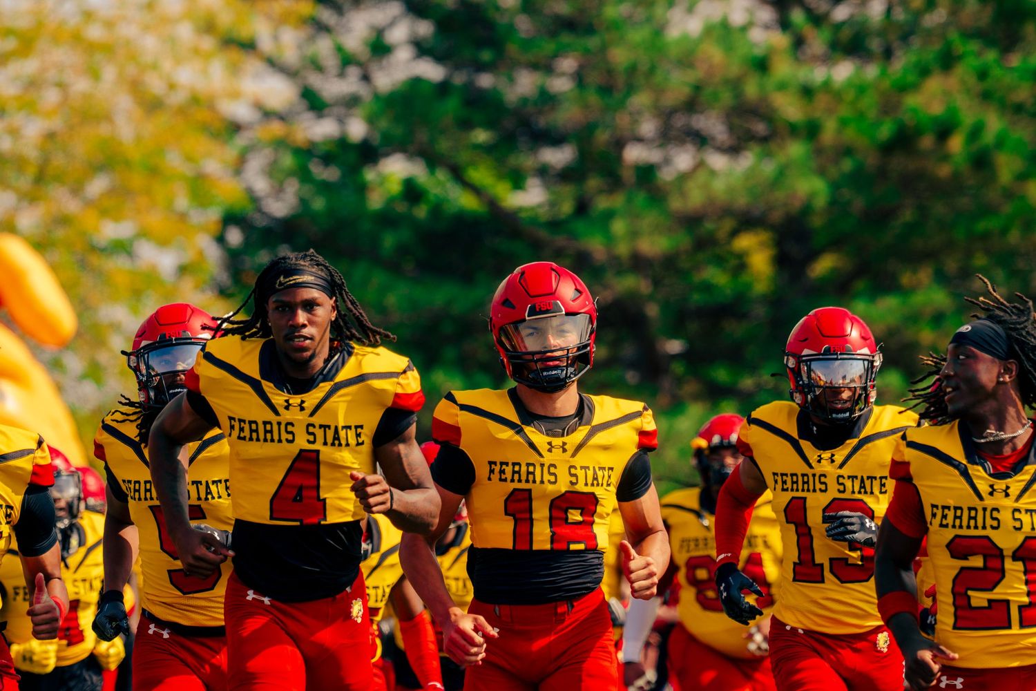 College football players in yellow jerseys and red helmets stand together on the sideline during a game.