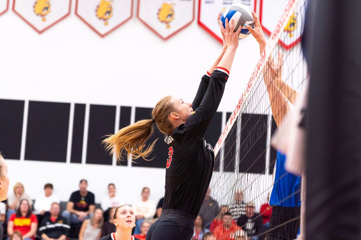 Female volleyball player in a black jersey leaps at the net to block a spike during a indoor match while spectators watch from the stands.