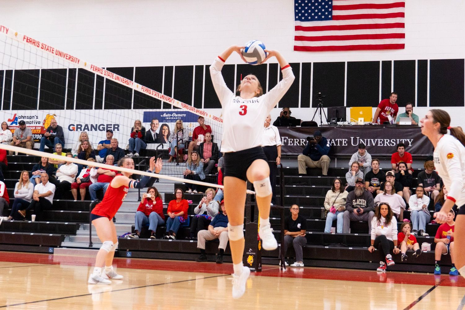 Volleyball player in a white number 3 jersey leaps to spike the ball during a college match as teammates and fans watch from the stands.