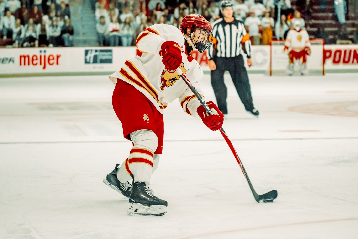 A hockey player in a red and gold uniform winds up to take a slap shot on the ice while a referee watches in the background.
