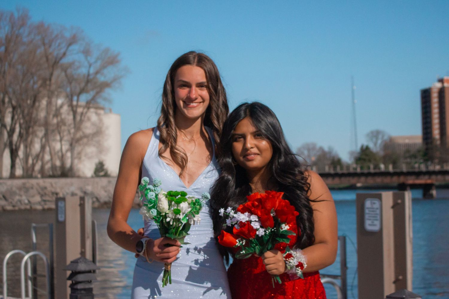 Two people holding colorful flower bouquets pose together by a waterfront railing.