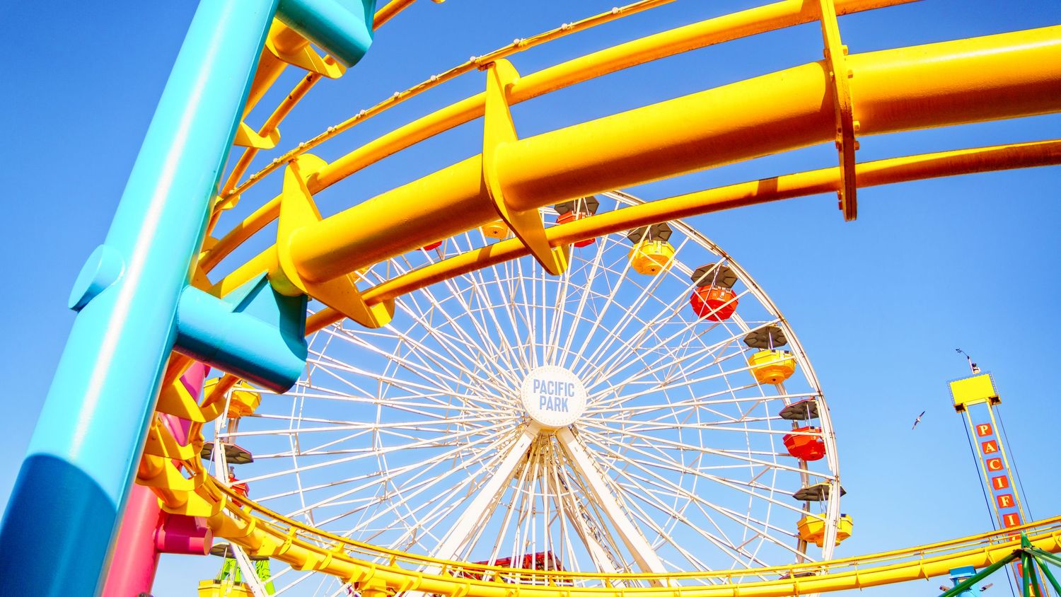 Carnival rides with bright blue and yellow metal structures frame a white Ferris wheel against the sky.