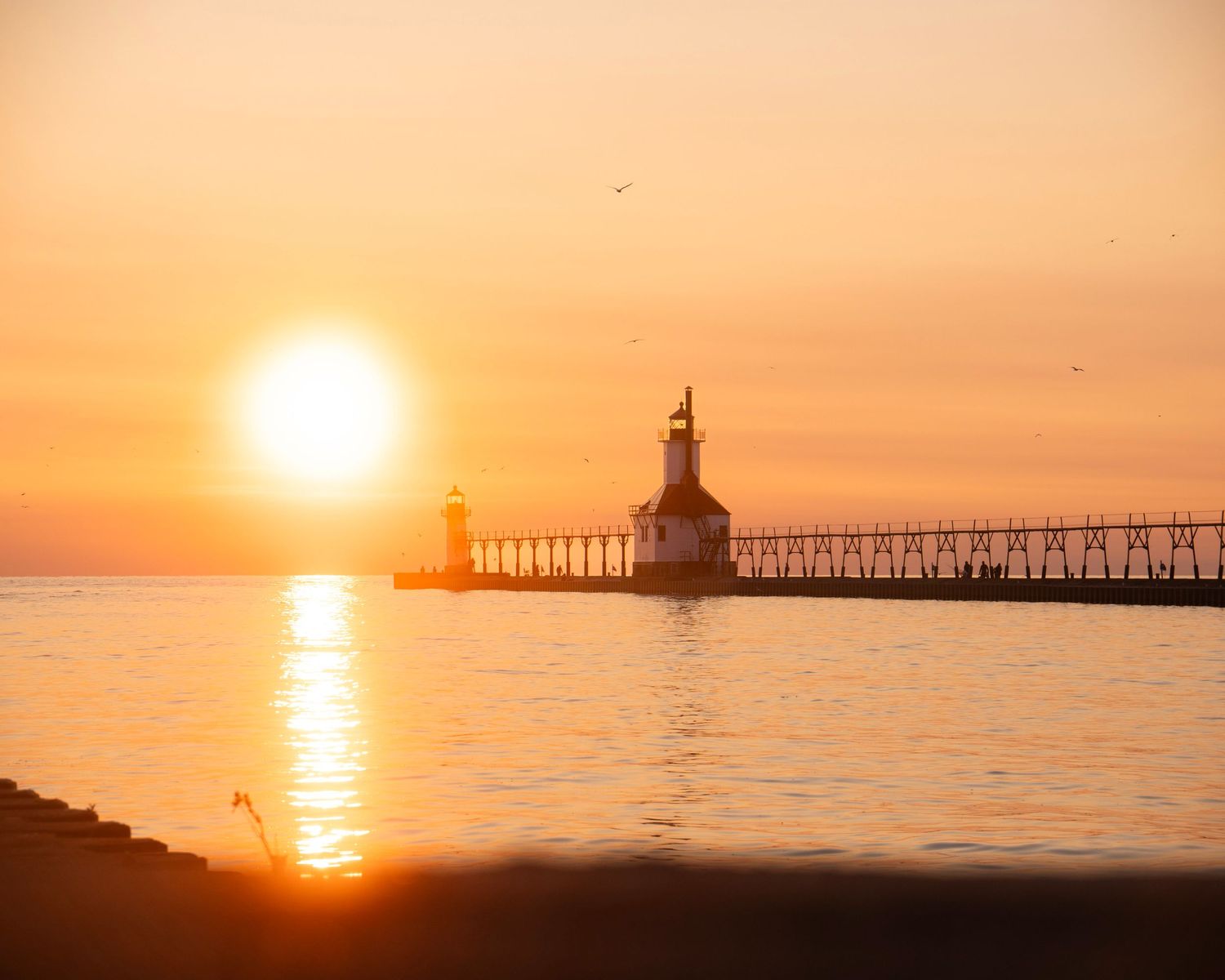 Lighthouse on Lake Michigan pier silhouetted against vibrant orange sunset with reflections on calm water.