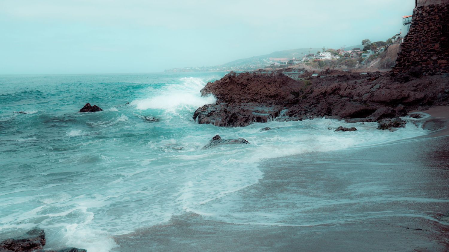 Misty turquoise waves crash against rocky coastal cliffs in a moody seascape.