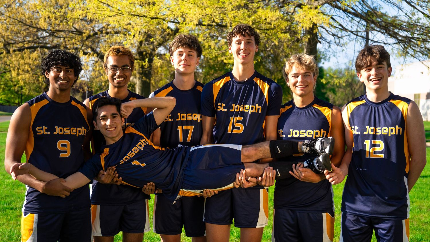 St Joseph volleyball team members in navy uniforms posed outdoors with fall foliage.