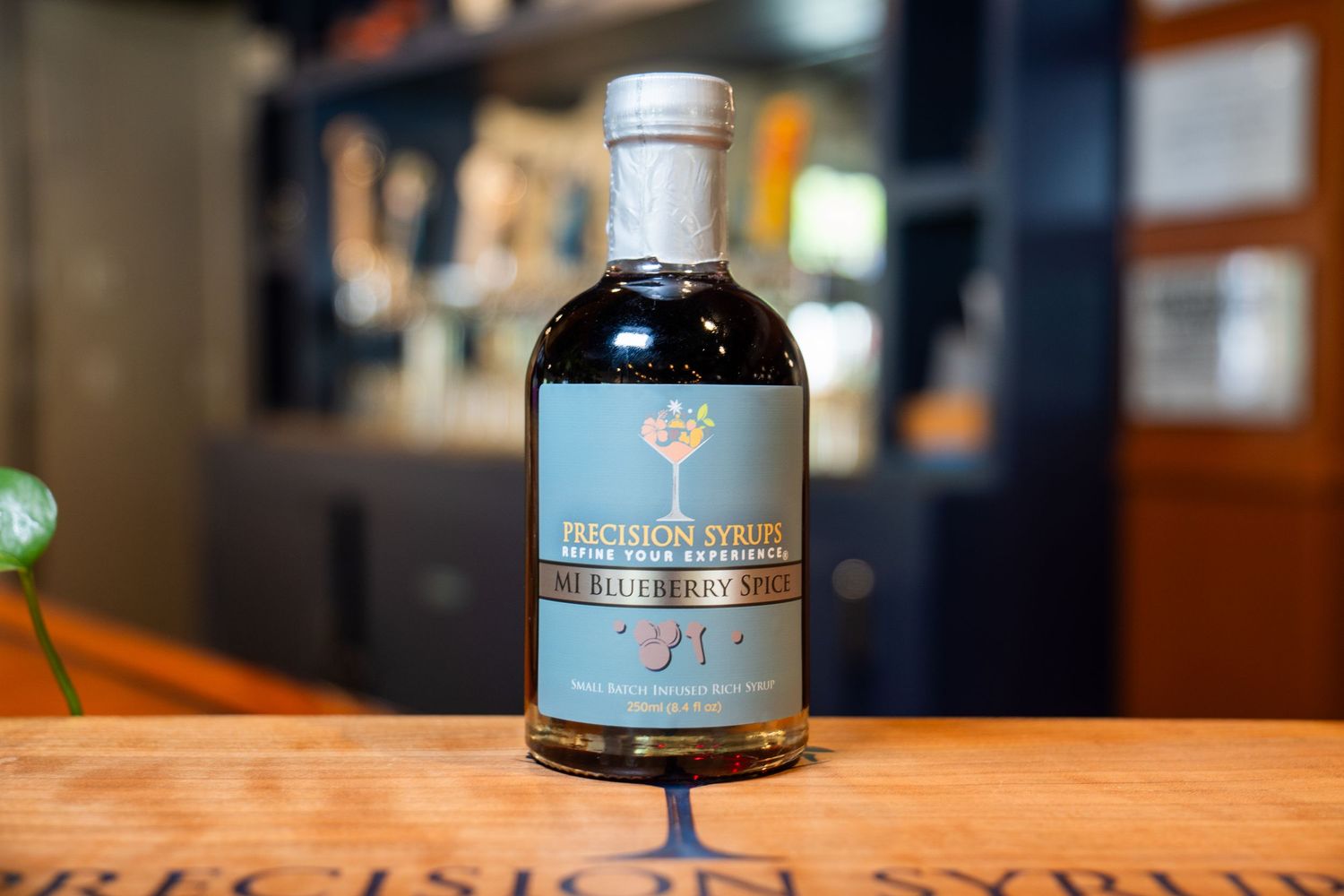 Dark bottle of craft cocktail syrup with blue label against blurred bar background.