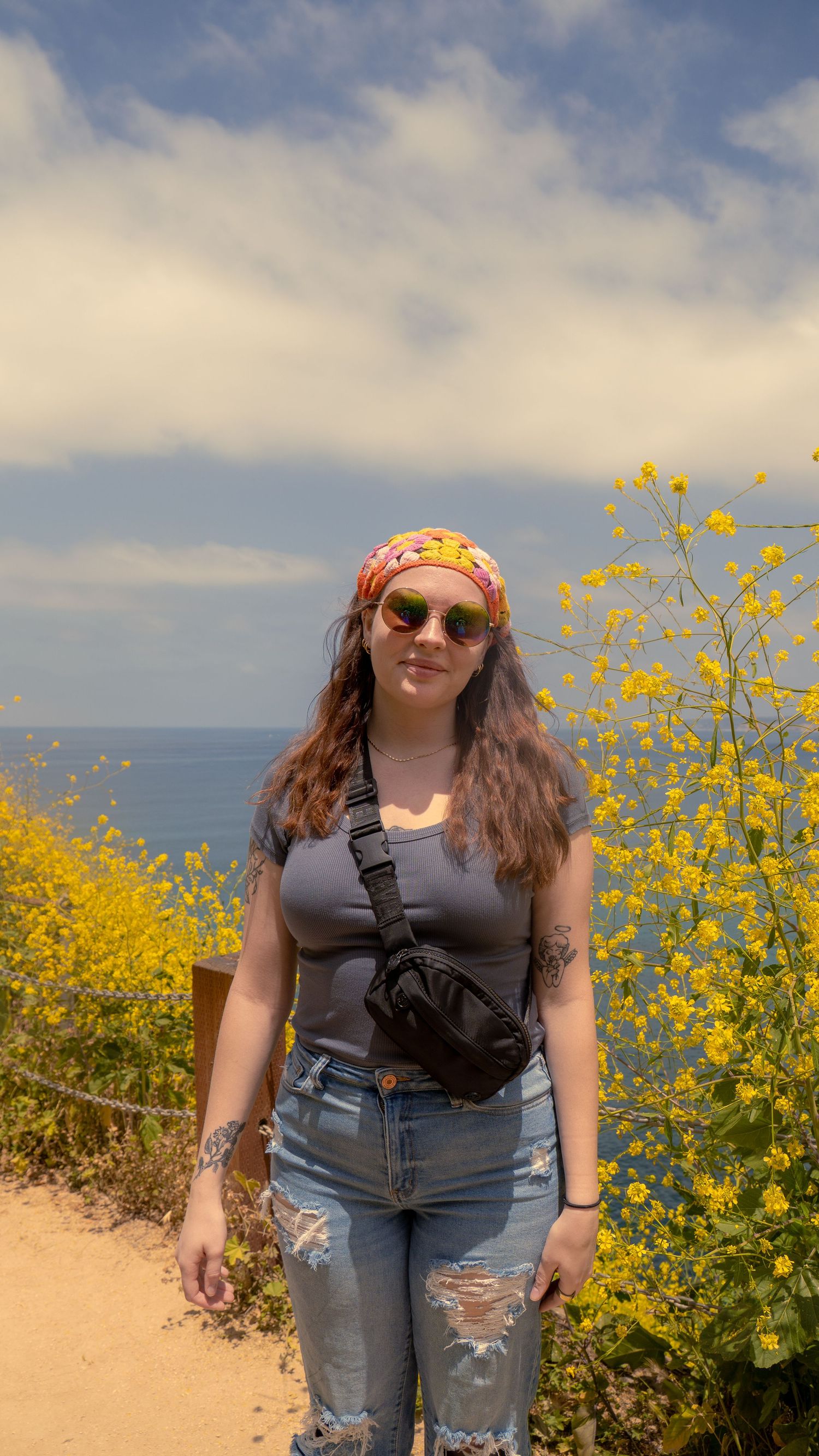 Hiker with fanny pack and bandana poses on coastal trail surrounded by yellow wildflowers.