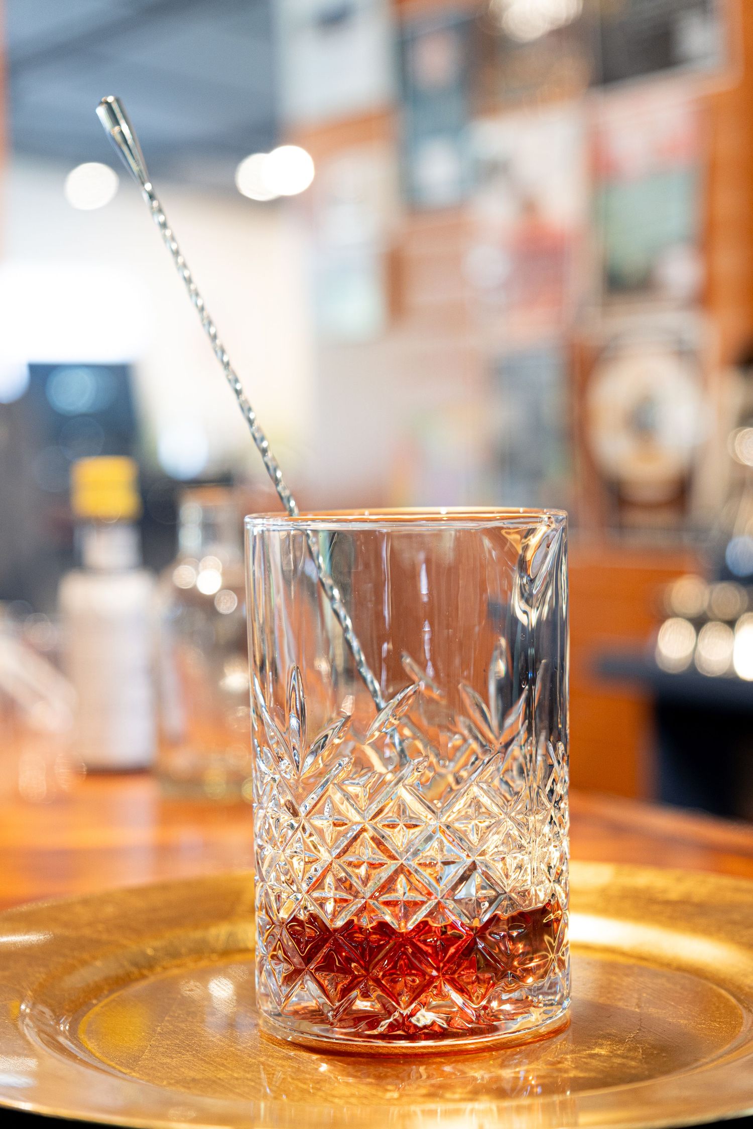 Cut crystal mixing glass with red cocktail syrup and striped straw on golden plate.