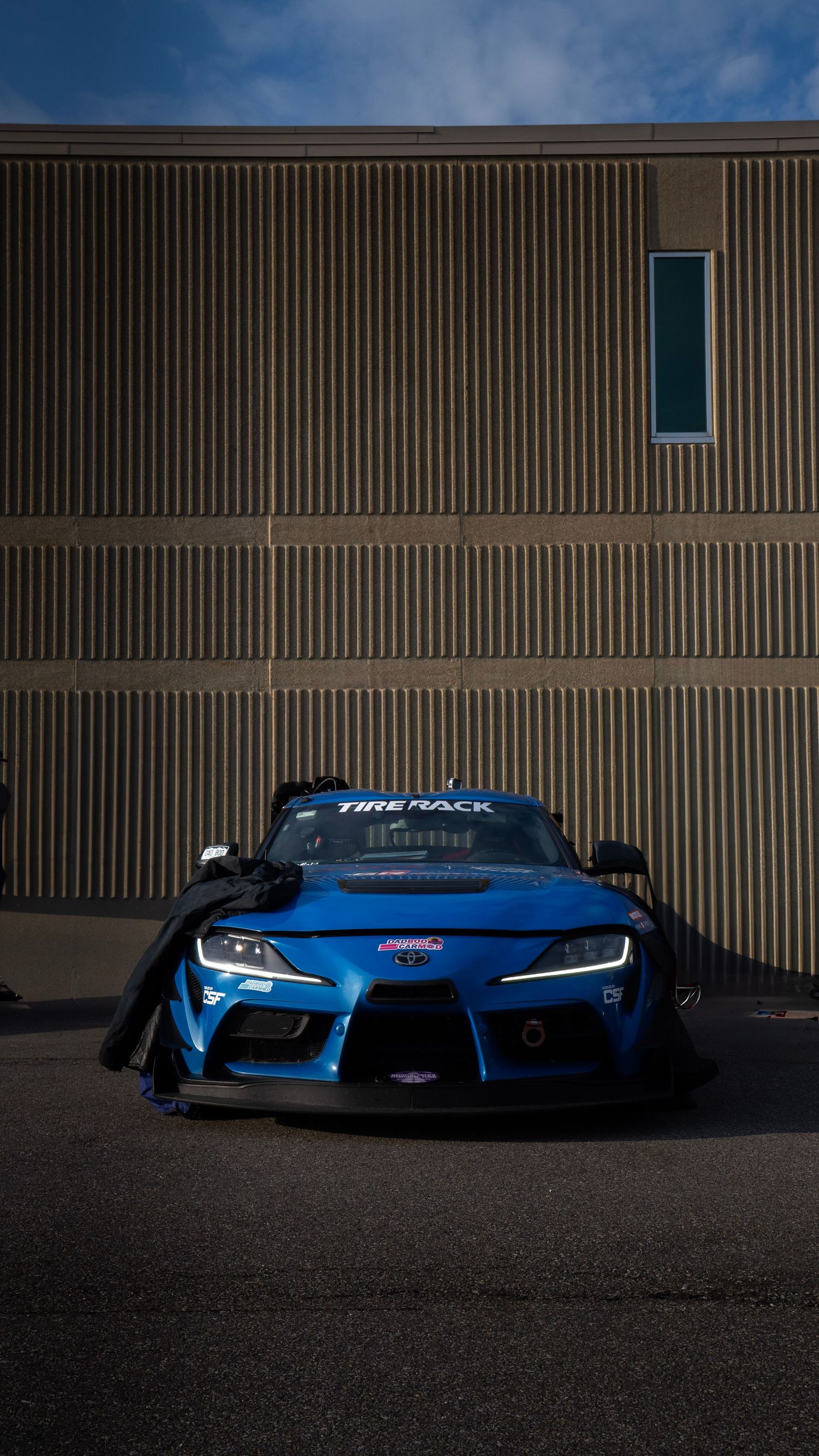 Blue Toyota Supra with aggressive body kit parked against industrial building.