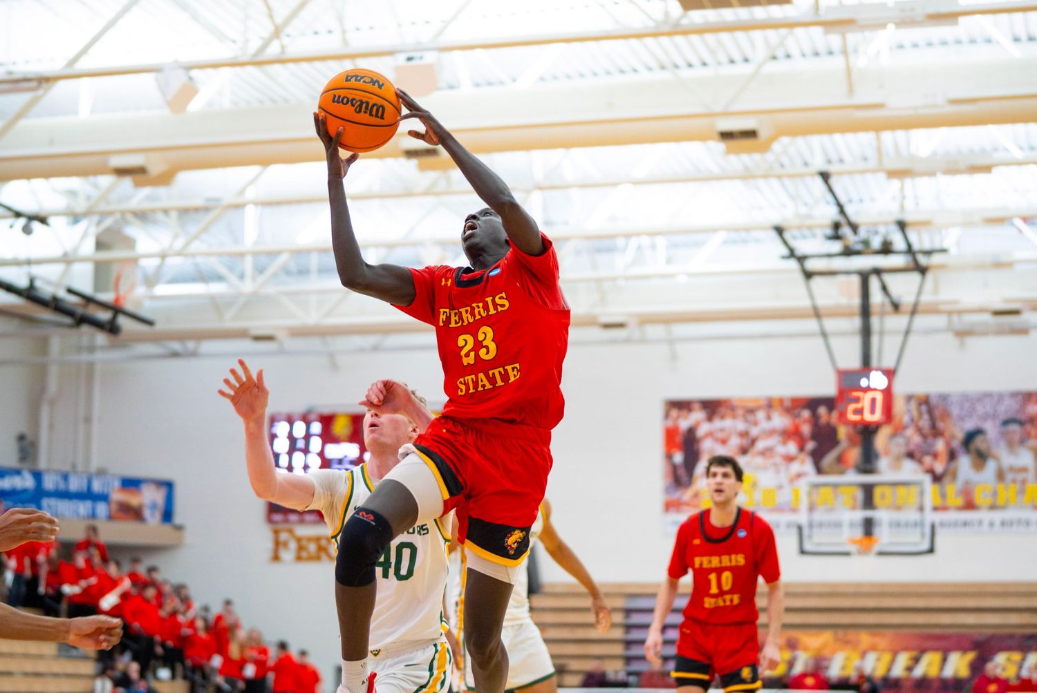Basketball player in red jersey leaps high for a shot during indoor game while defender attempts to block.