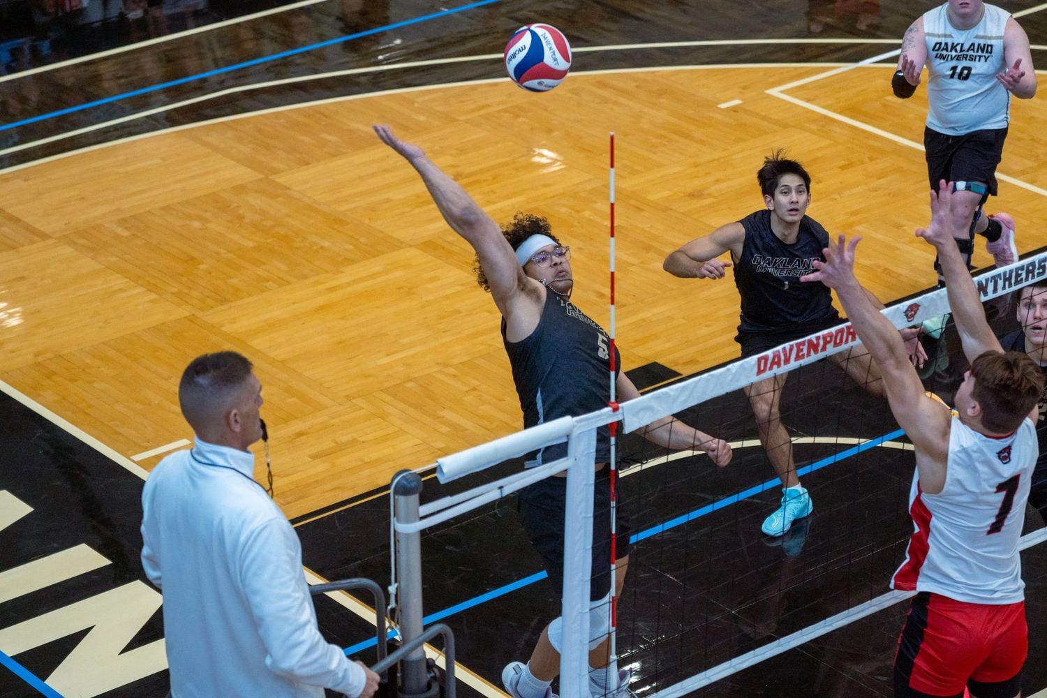 A volleyball player jumps to spike the ball during an intense indoor match on a blue court.