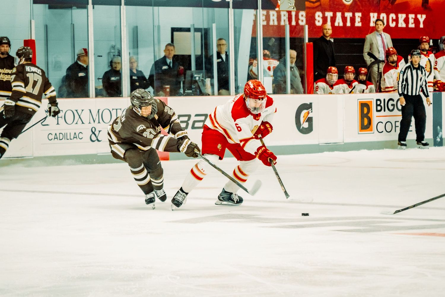 Two college hockey players, one in a white and red uniform and one in a dark uniform, battle for the puck on the ice during a game.