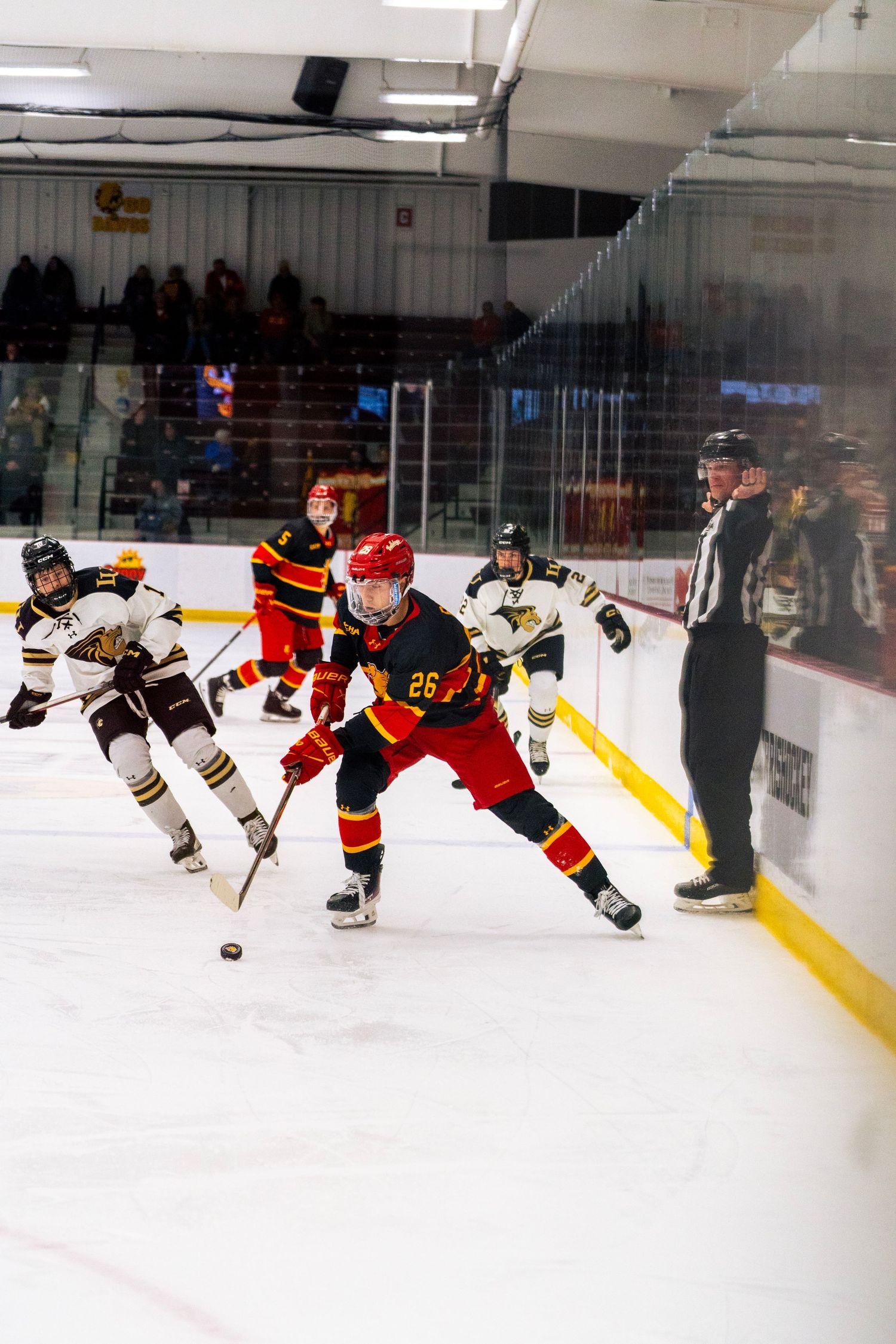 Hockey player in red and black jersey number 26 skates after the puck while opponents in white and a referee follow along the boards.