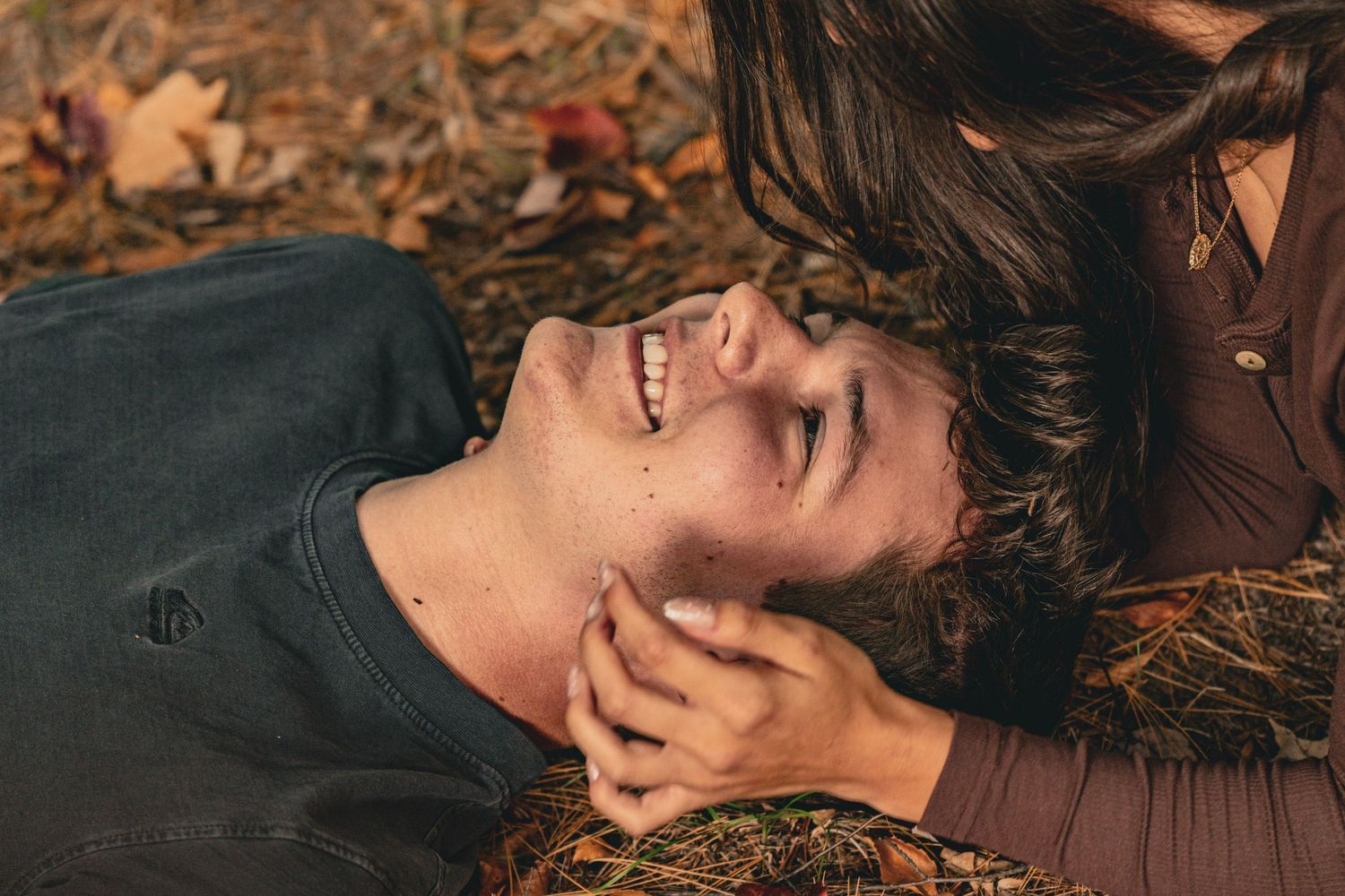Smiling young man lying on autumn leaves while a woman with long dark hair leans over and gently touches his face.