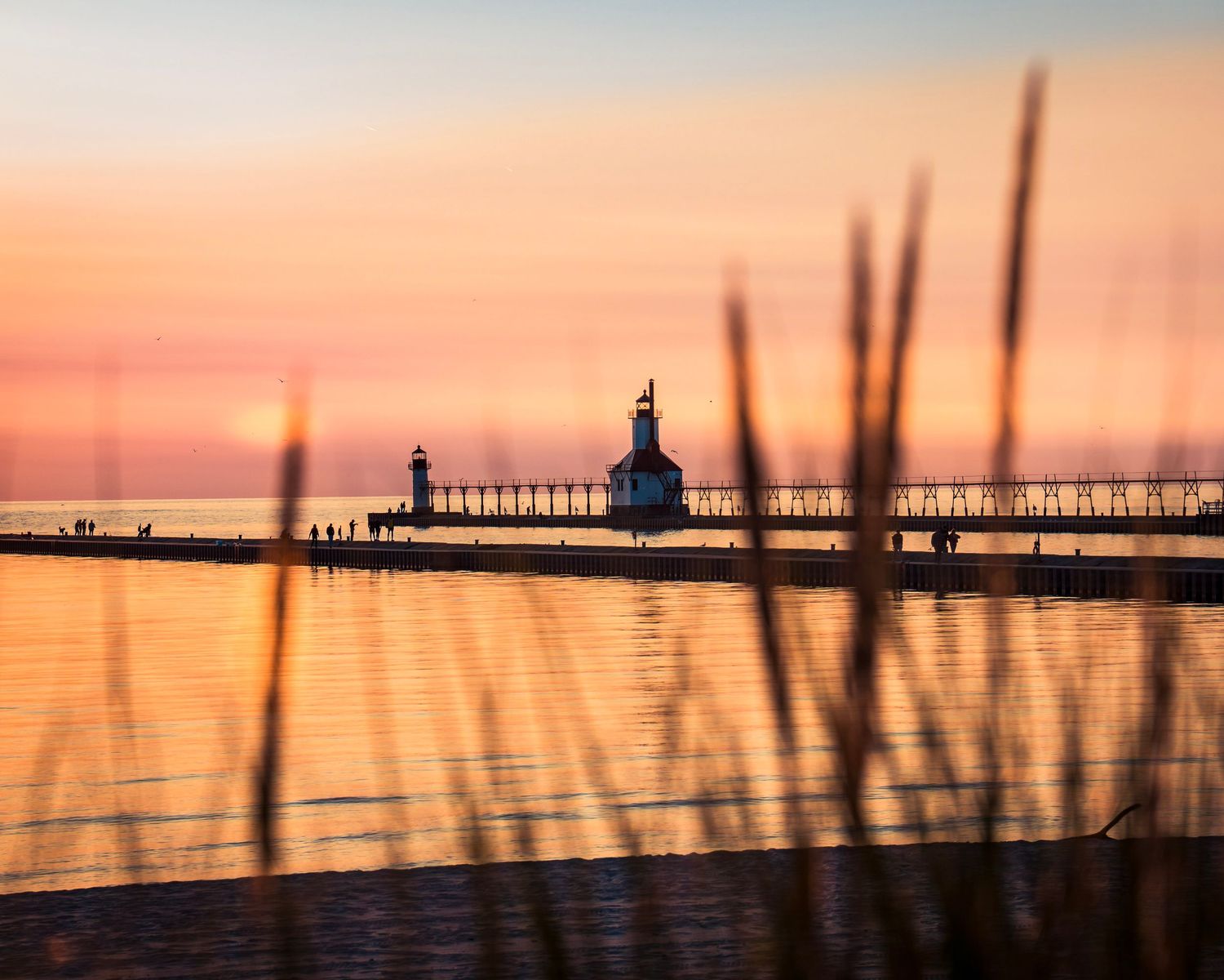 Beach grass silhouettes frame distant pier and lighthouse at dusk.