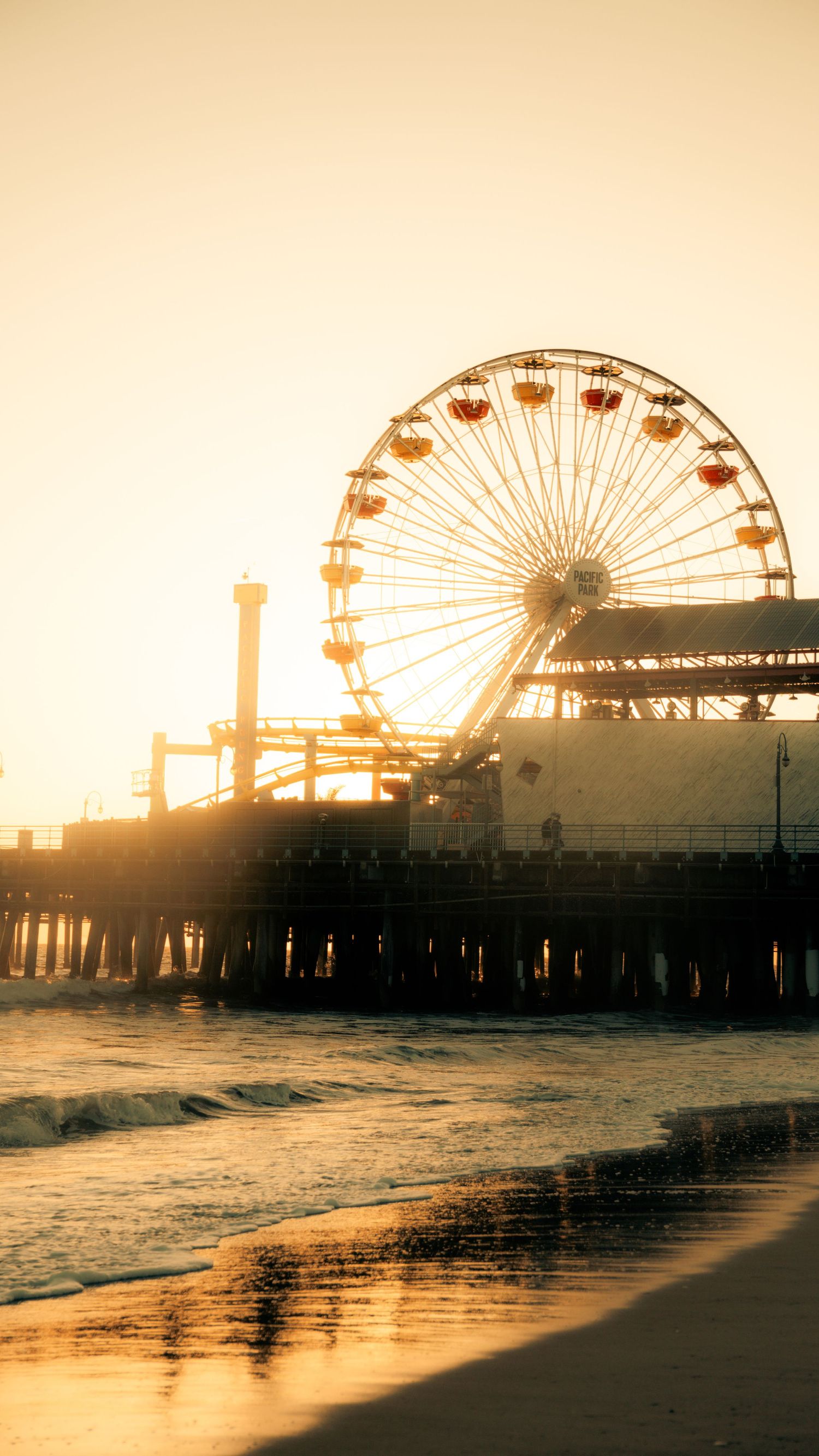 Ferris wheel silhouetted against golden sunset at Santa Monica Pier with waves reflecting on beach.