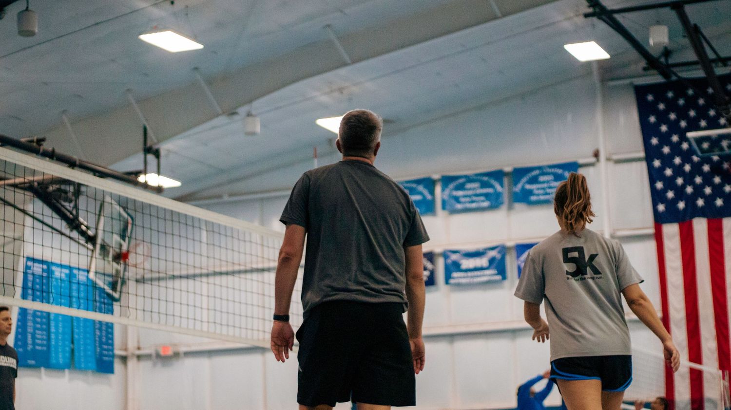 Two players standing by volleyball net in gymnasium with American flag and blue banners.