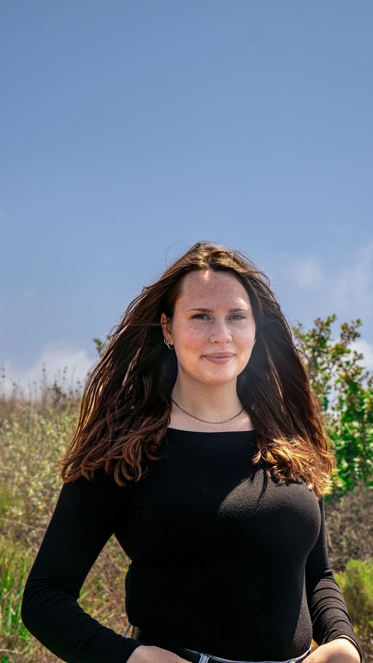 Portrait of person in black long-sleeve top smiling against blue sky and natural landscape.