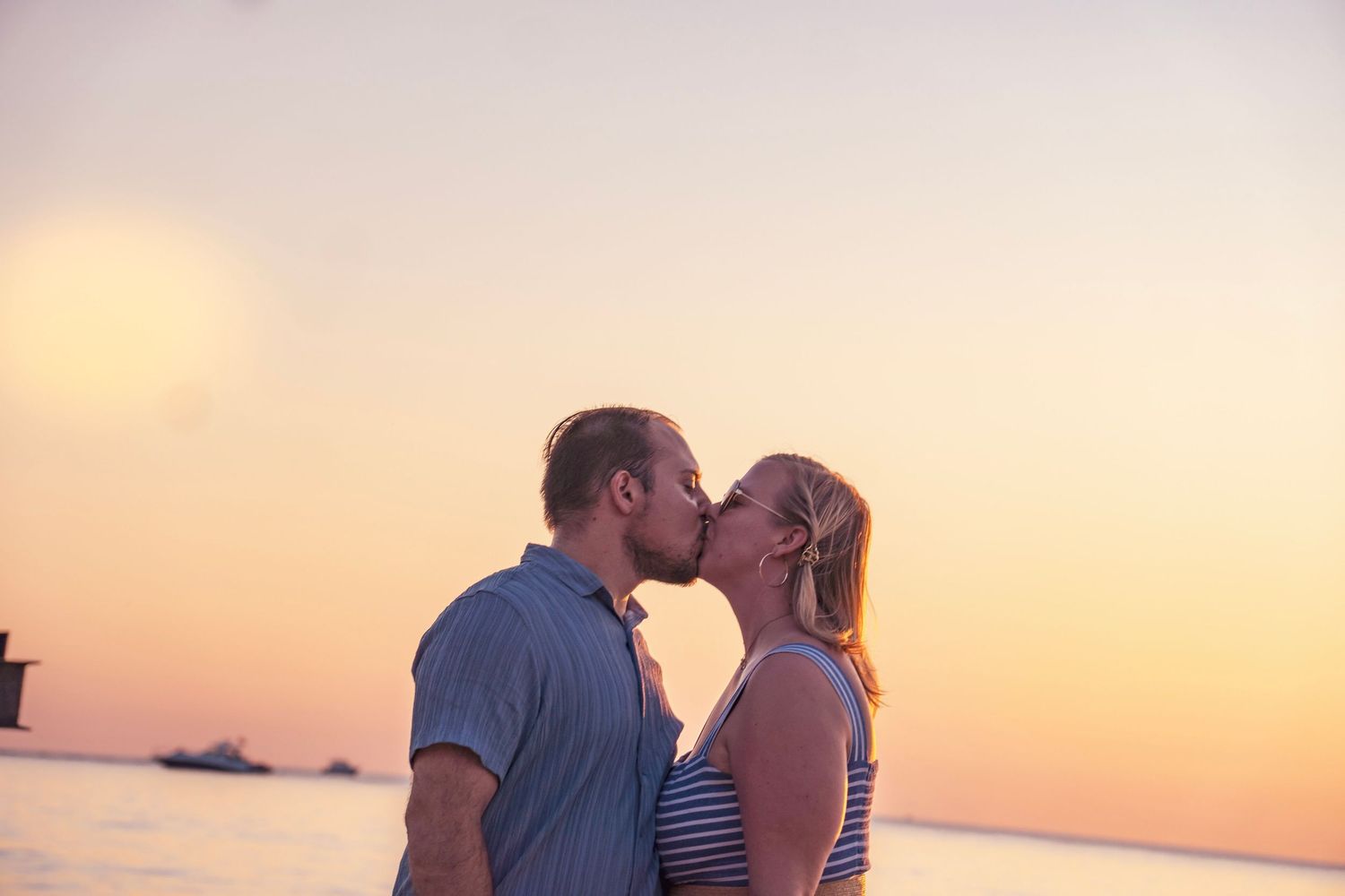 A couple shares an intimate kiss against a beautiful orange sunset sky over calm ocean waters.