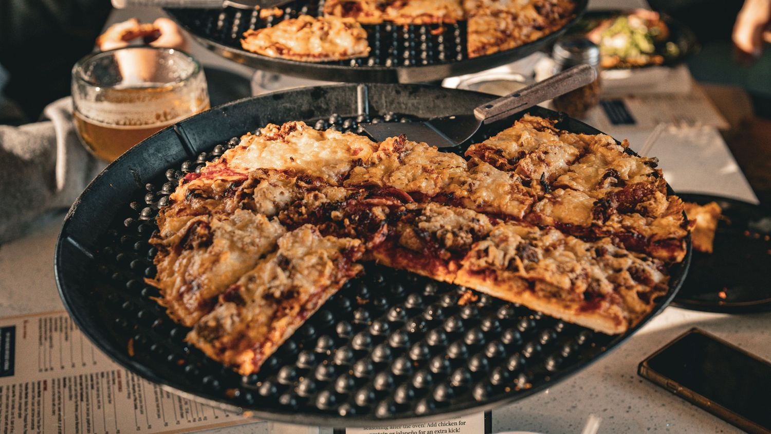 Close-up view of sliced pepperoni pizza on a black perforated pan with assorted toppings and melted cheese.
