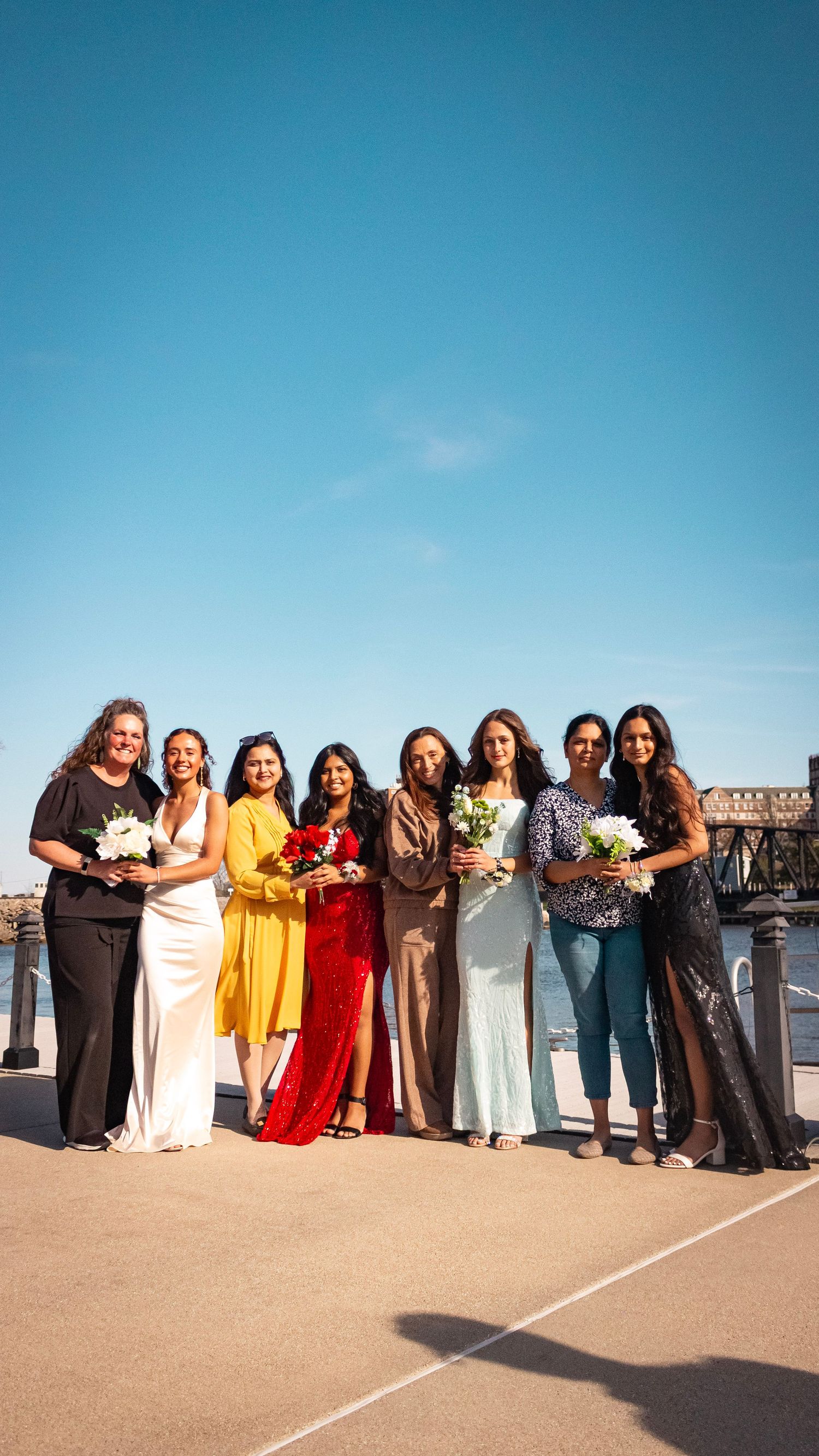 A group of people in colorful formal attire stand together in a line outdoors against a bright blue sky.