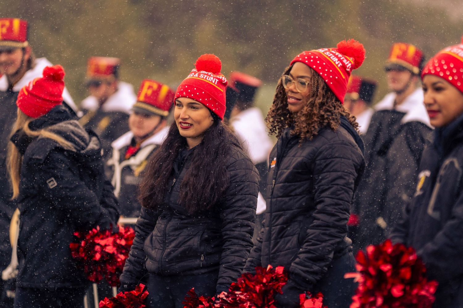 A row of cheerleaders wearing red winter hats and black coats perform at a sporting event.