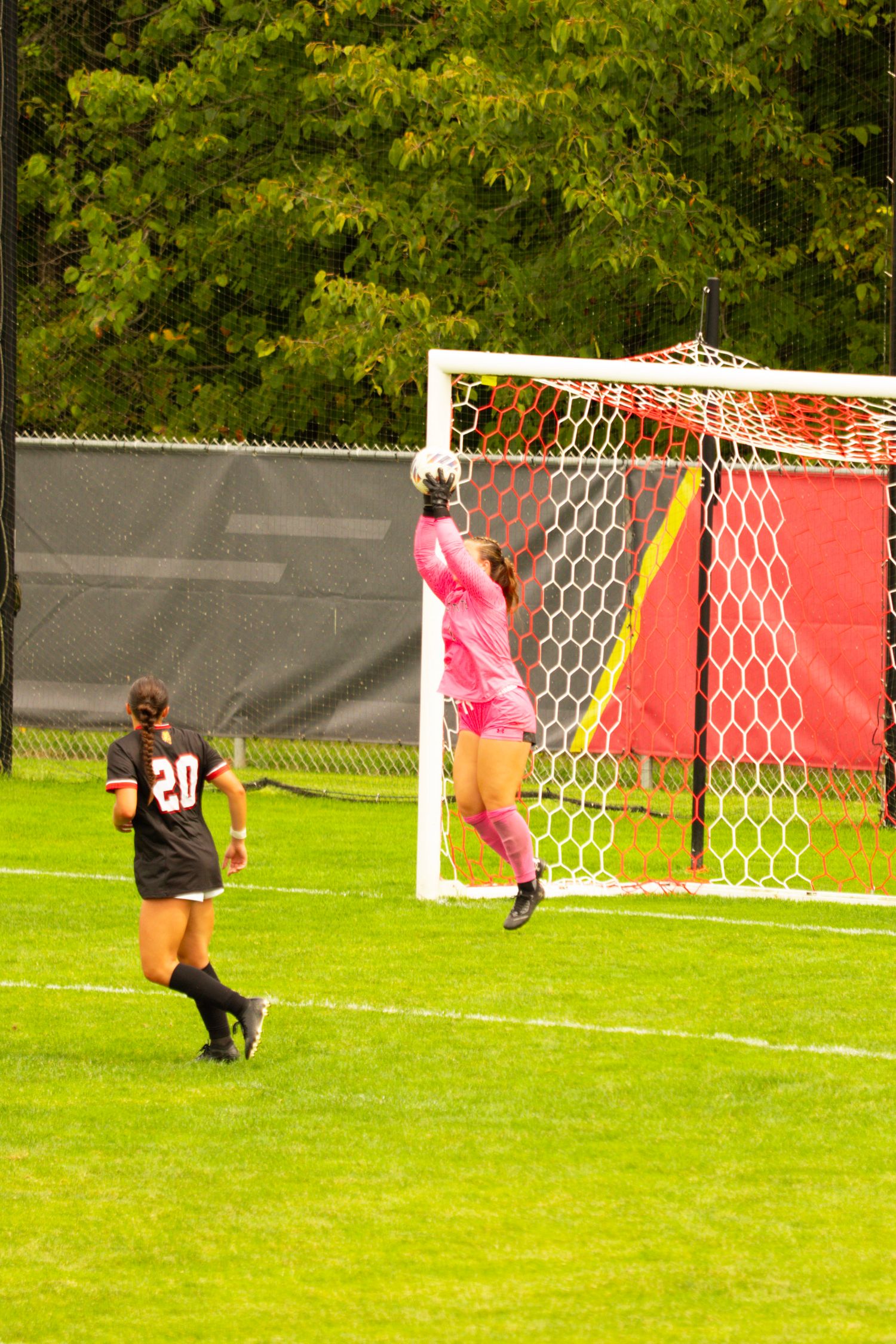 Goalkeeper in a pink uniform leaps to catch a soccer ball in front of the goal while a teammate wearing number 20 looks on.