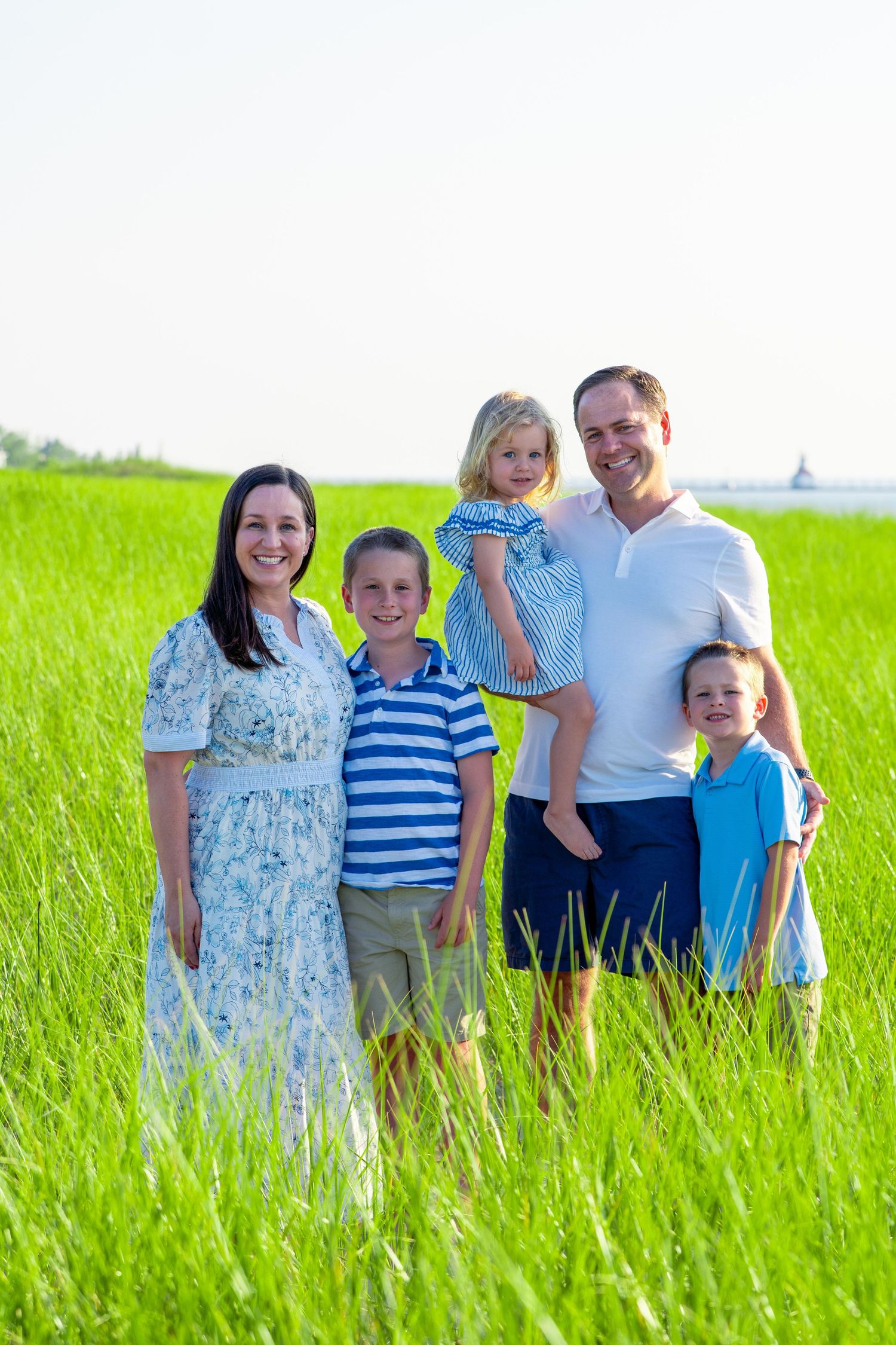 Smiling family of five, two adults and three children, posing together in a bright green grassy field on a sunny day.