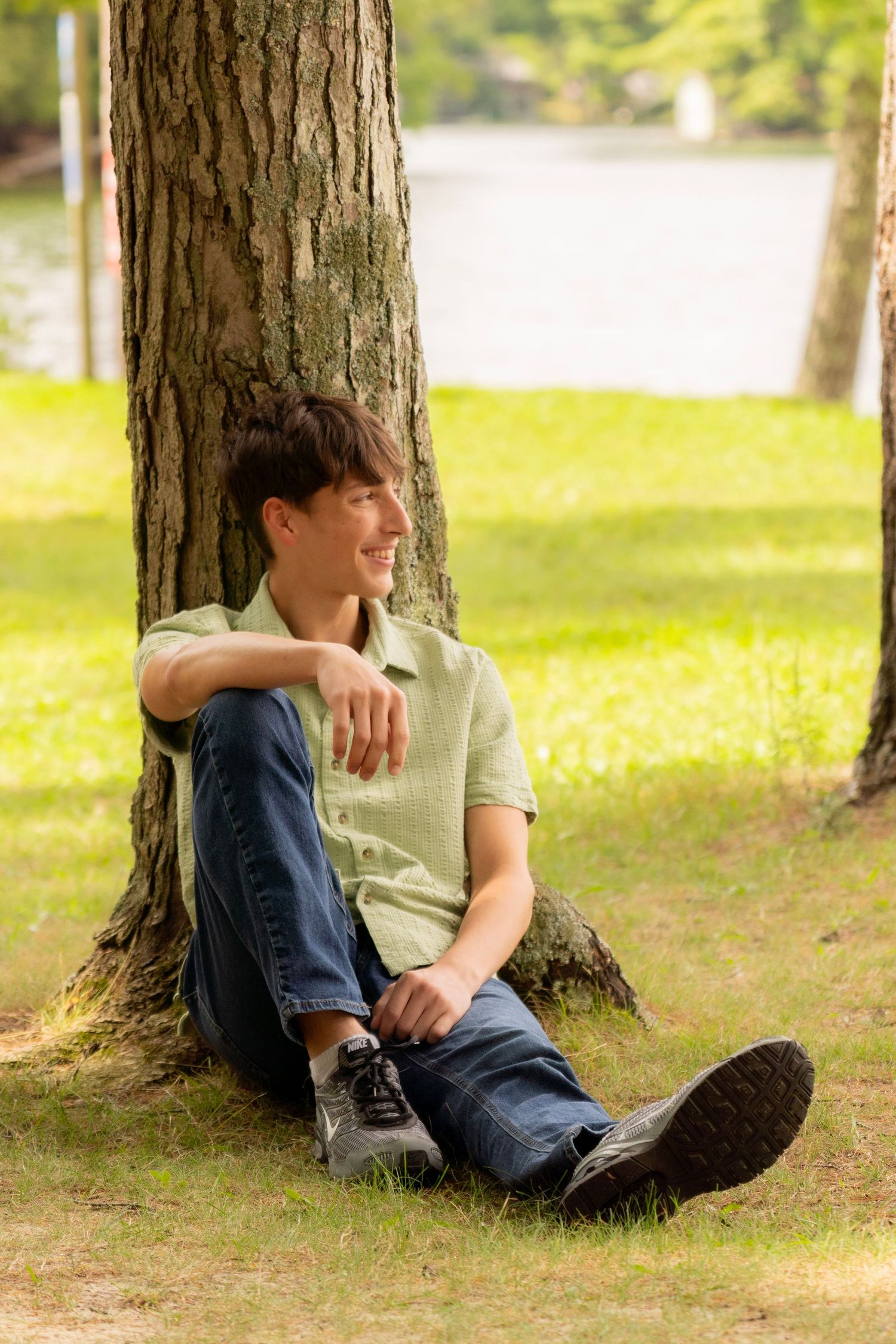 Smiling young man in a green button-up shirt and jeans sits against a tree trunk on sunlit grass near a lake.