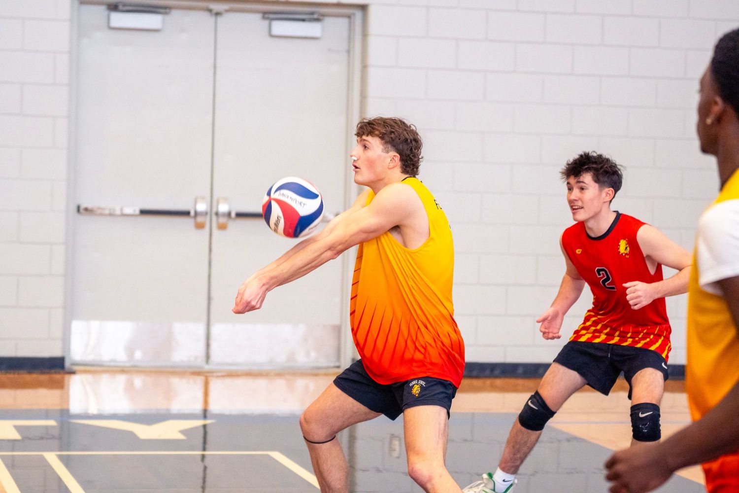 Players in orange and red jerseys compete in an indoor volleyball match, diving and jumping for the ball.