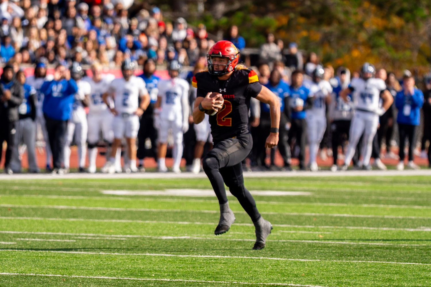 College football player in red helmet and black uniform runs with ball during game on sunny autumn day.