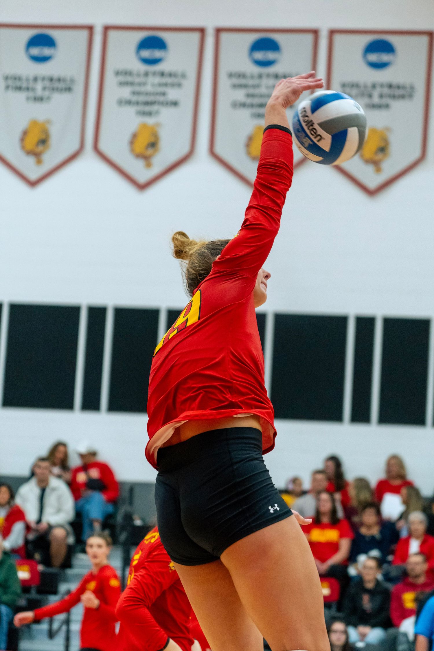 Volleyball players in red uniforms jump high at the net during an intense indoor match with spectators watching.