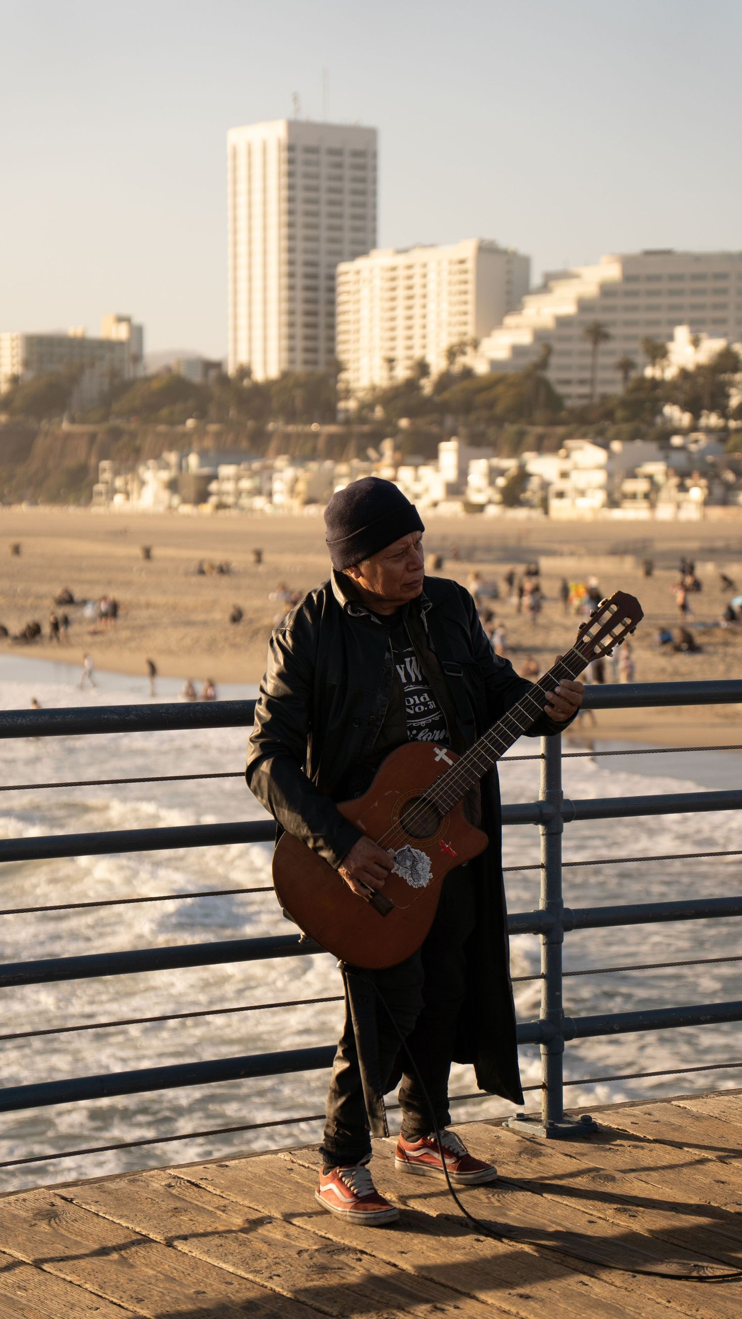 Street musician playing guitar on oceanfront boardwalk with city buildings in background.