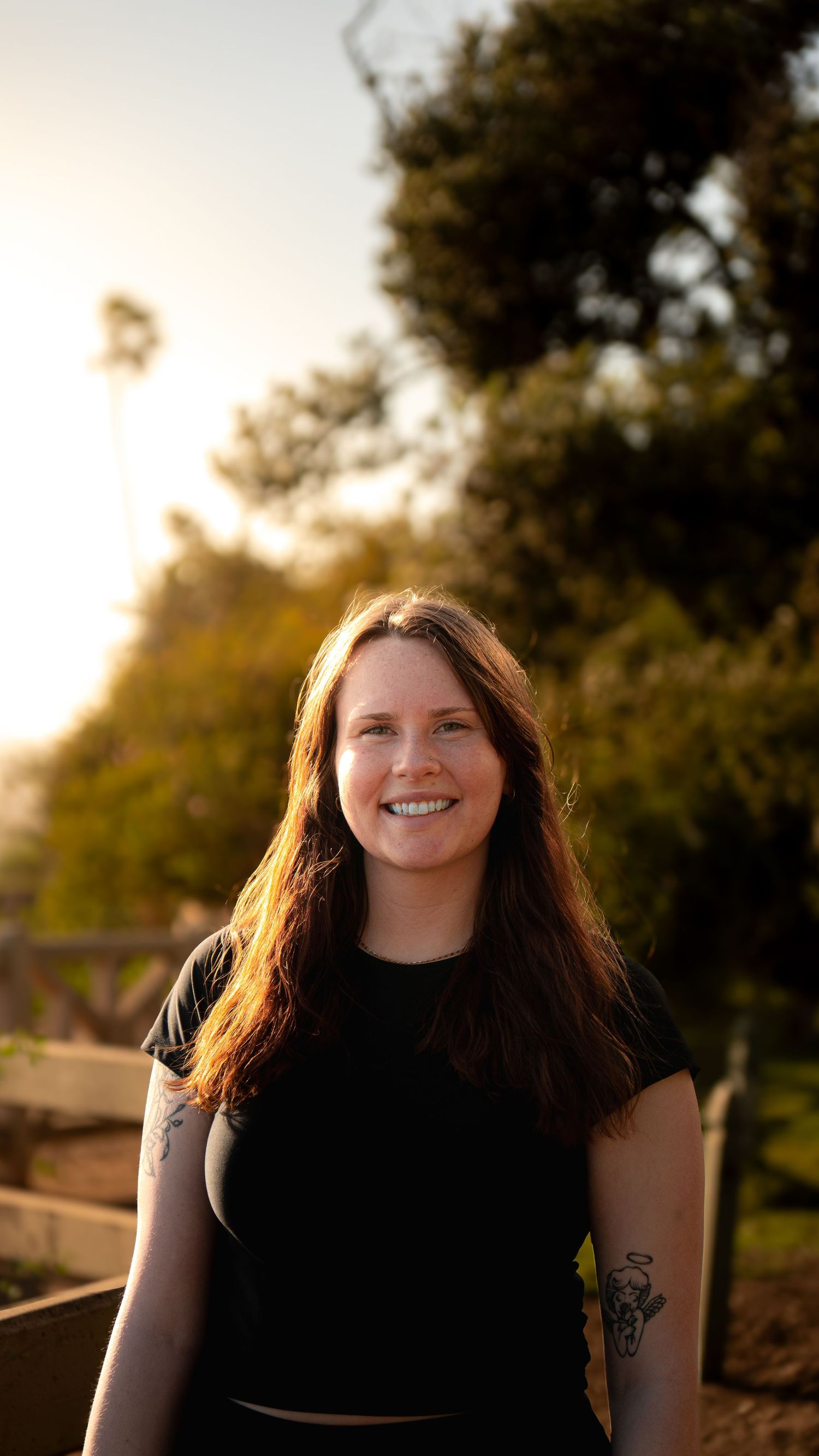Person in black top poses outdoors during sunset with trees in background.