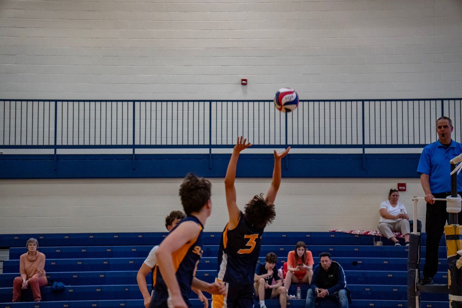 Two volleyball players competing for the ball during an indoor game with blue bleachers in the background.