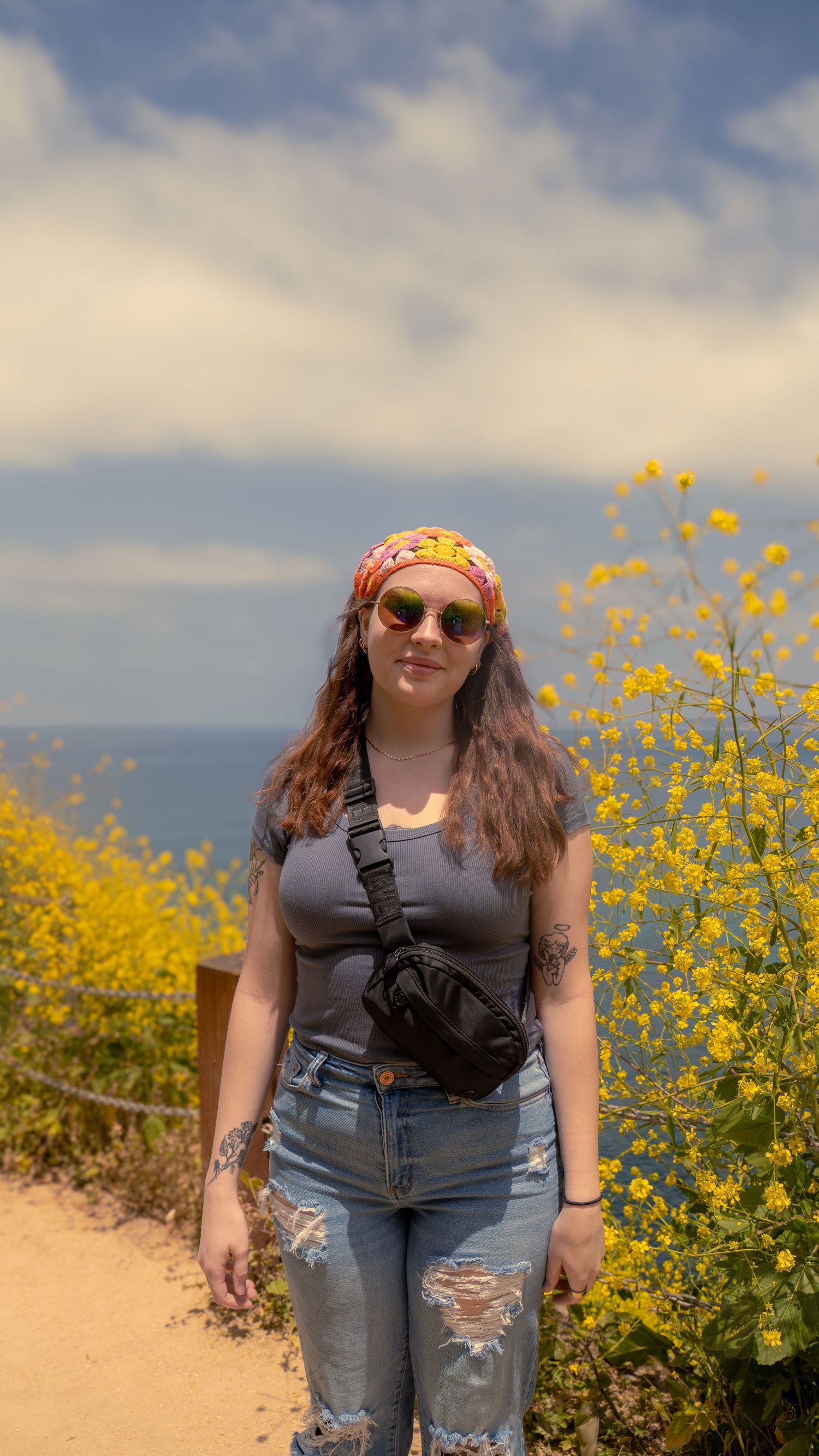 Person wearing sunglasses and bandana poses on hiking trail surrounded by vibrant yellow wildflowers on coastal path.