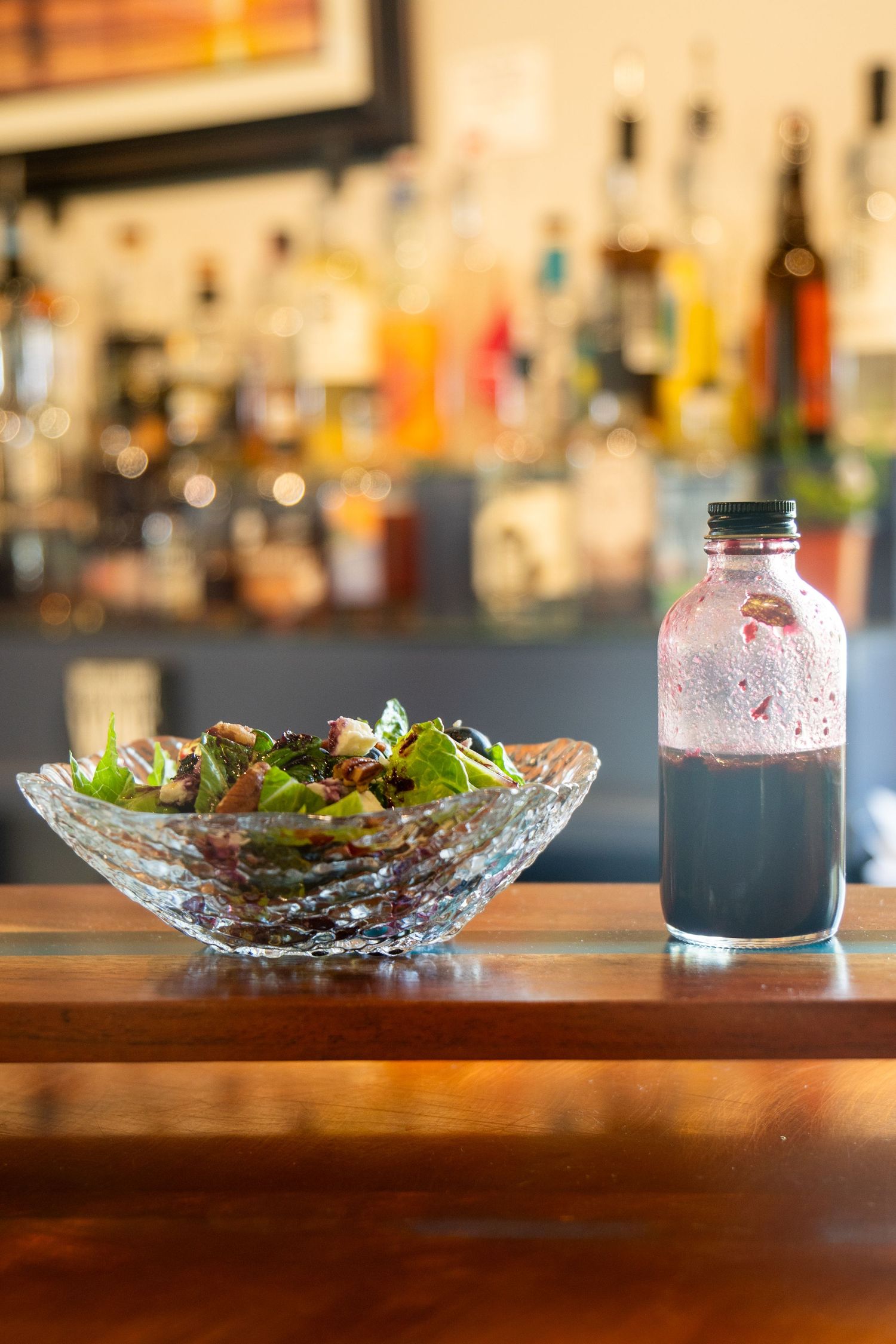 Green salad in cut crystal bowl next to dark bottled dressing on bar counter.