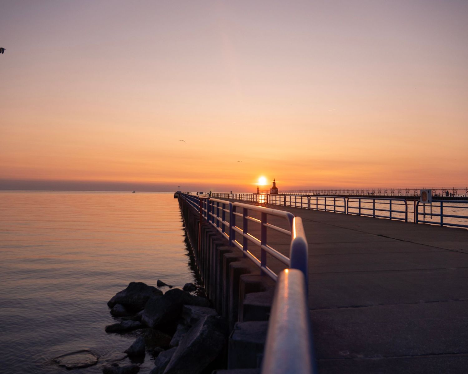 Metal railing leads to lighthouse at end of pier during peaceful sunset.